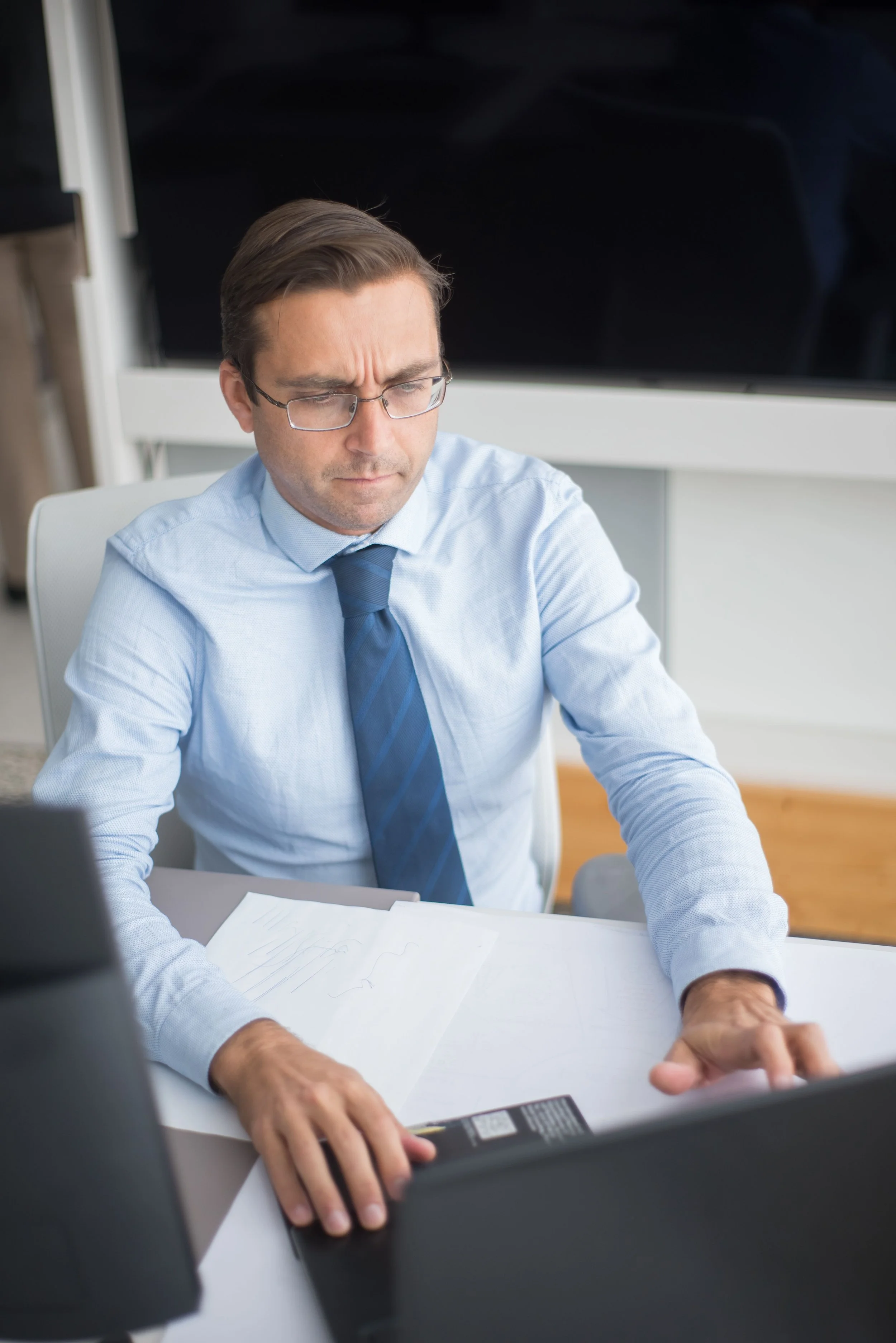 Homme d'affaires portant une chemise bleue, une cravate, et des lunettes assis à un bureau, semblant réfléchir ou préoccupé, avec des documents et une calculatrice sur la table.