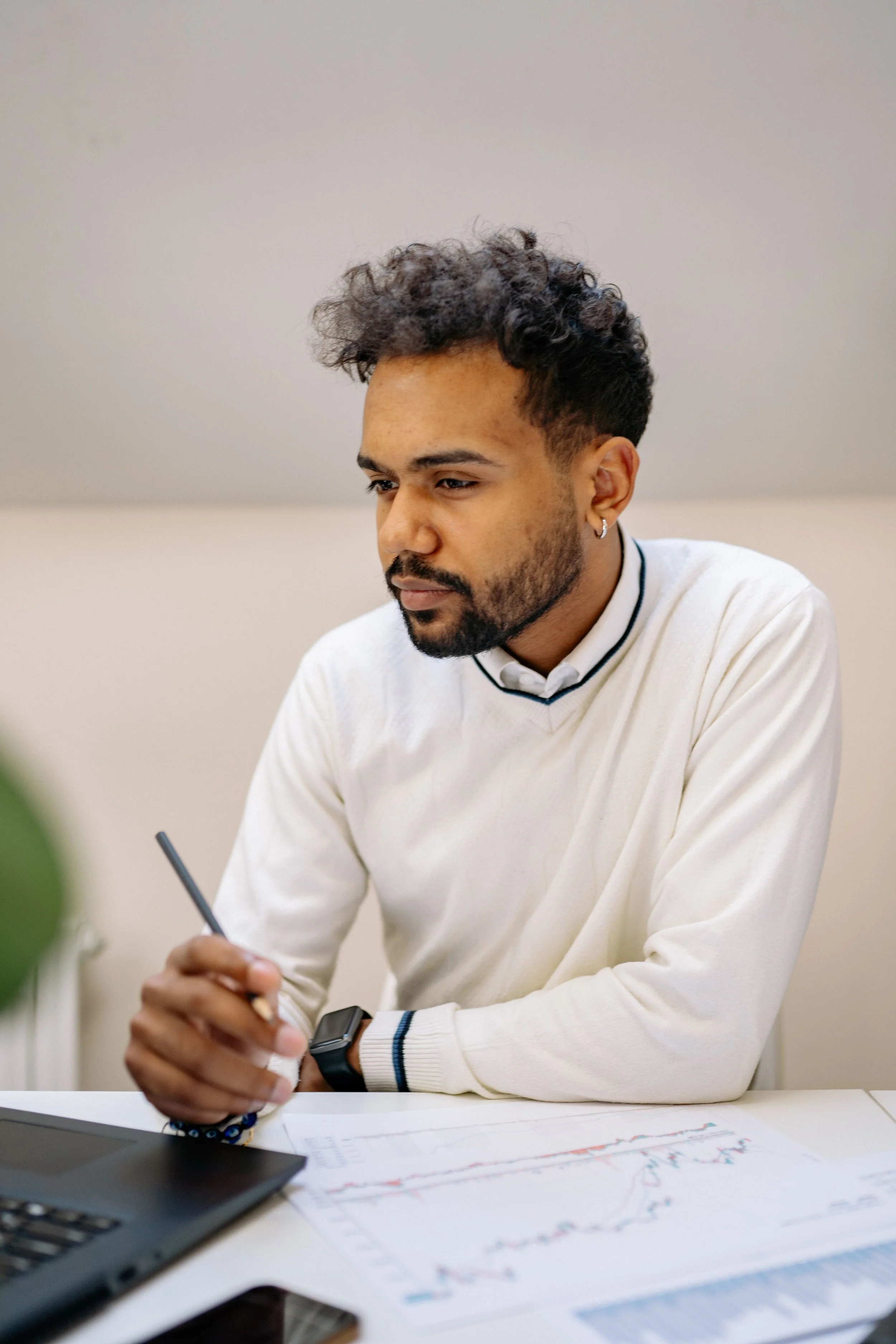 Un homme assis à une table avec des graphiques et documents, utilisant un stylo, portant une montre et une boucle d'oreille, dans un environnement intérieur.