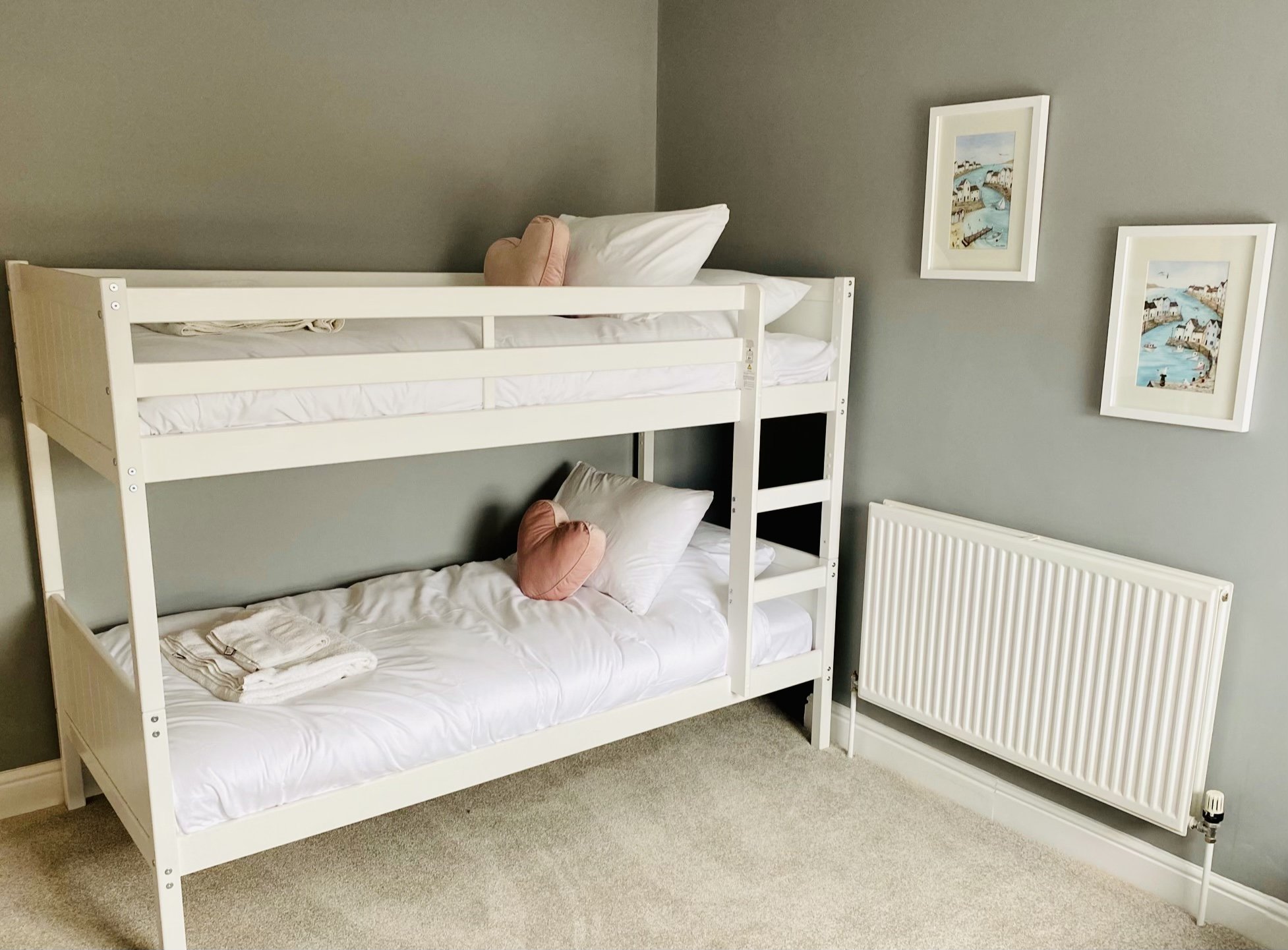 The childrens bunk bed room at Coast House, Cromer, with neatly made beds, pink heart-shaped pillows, a towel on the lower bed, and a stack of white towels, set against a grey wall with two framed seaside paintings, and a white radiator below.