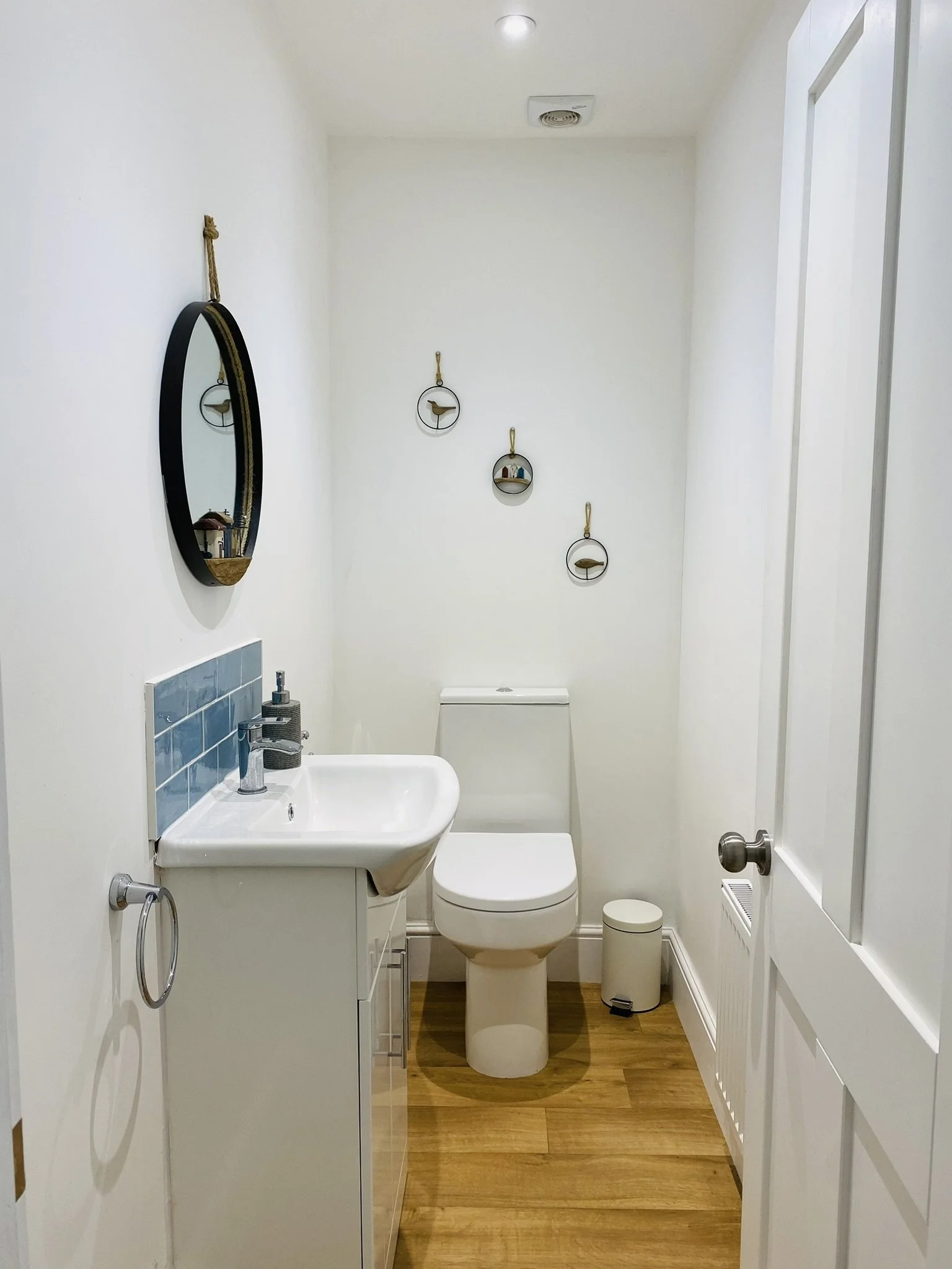 Downstairs toilet, coast themed, in Cromer, North Norfolk, a small, minimalistic bathroom with white walls and wooden flooring, featuring a white toilet, a small white sink with a mirror above, and wall decorations with nautical themes.
