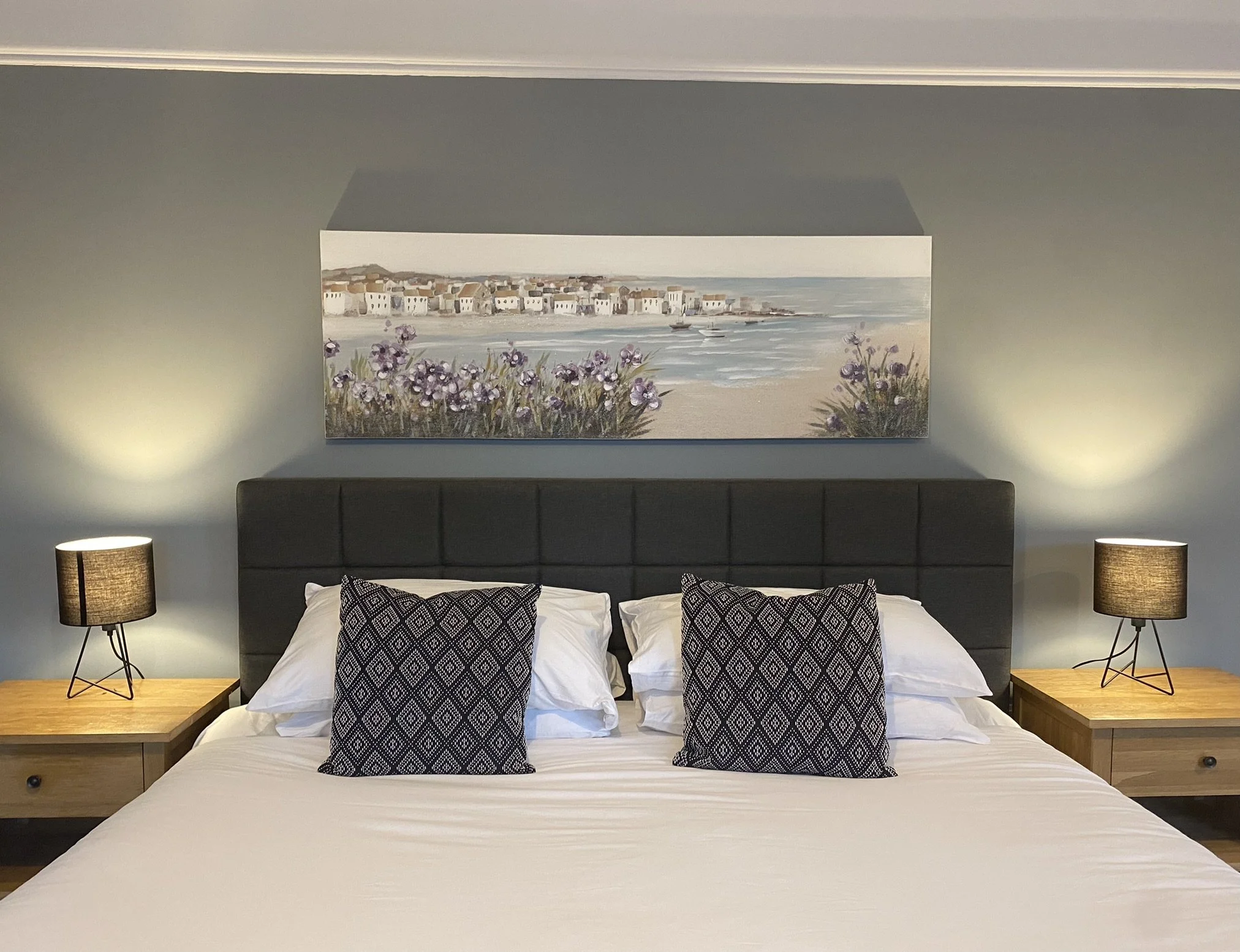 A bedroom with a bed that has white sheets, decorative black and white patterned pillows, and a padded dark grey headboard. There are two wooden nightstands with black table lamps on each side and a framed landscape painting of a coastal scene with purple flowers on the wall above the bed.