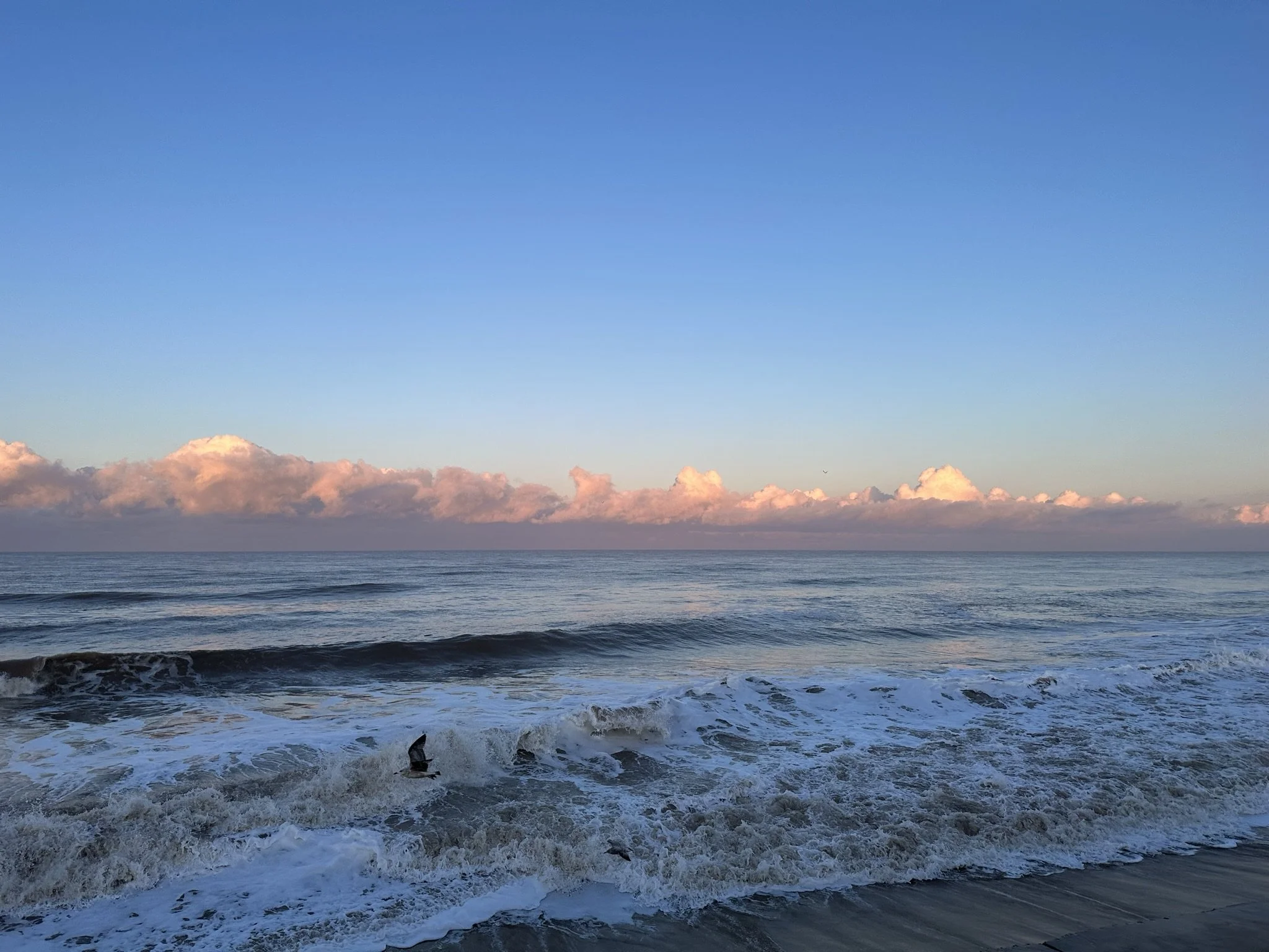 A calm ocean scene at Bacton beach, North Norfolk, with gentle waves and pink-tinged clouds on the horizon under a clear blue sky.