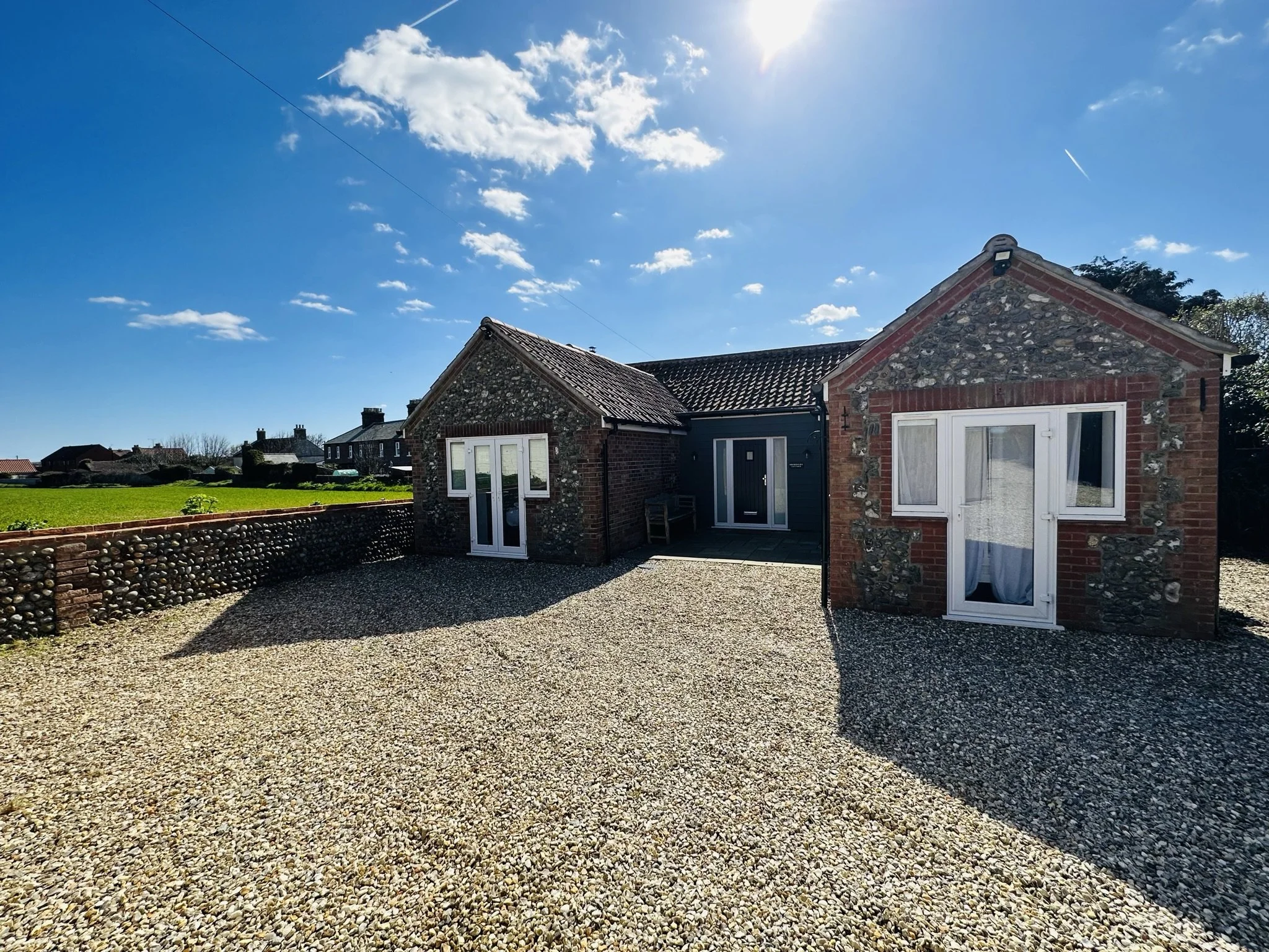 Bromholme Cottage, Bacton, North Norfolk, with large  gravel driveway for four cars in front of a flint frontcottage, under a blue sky with clouds.