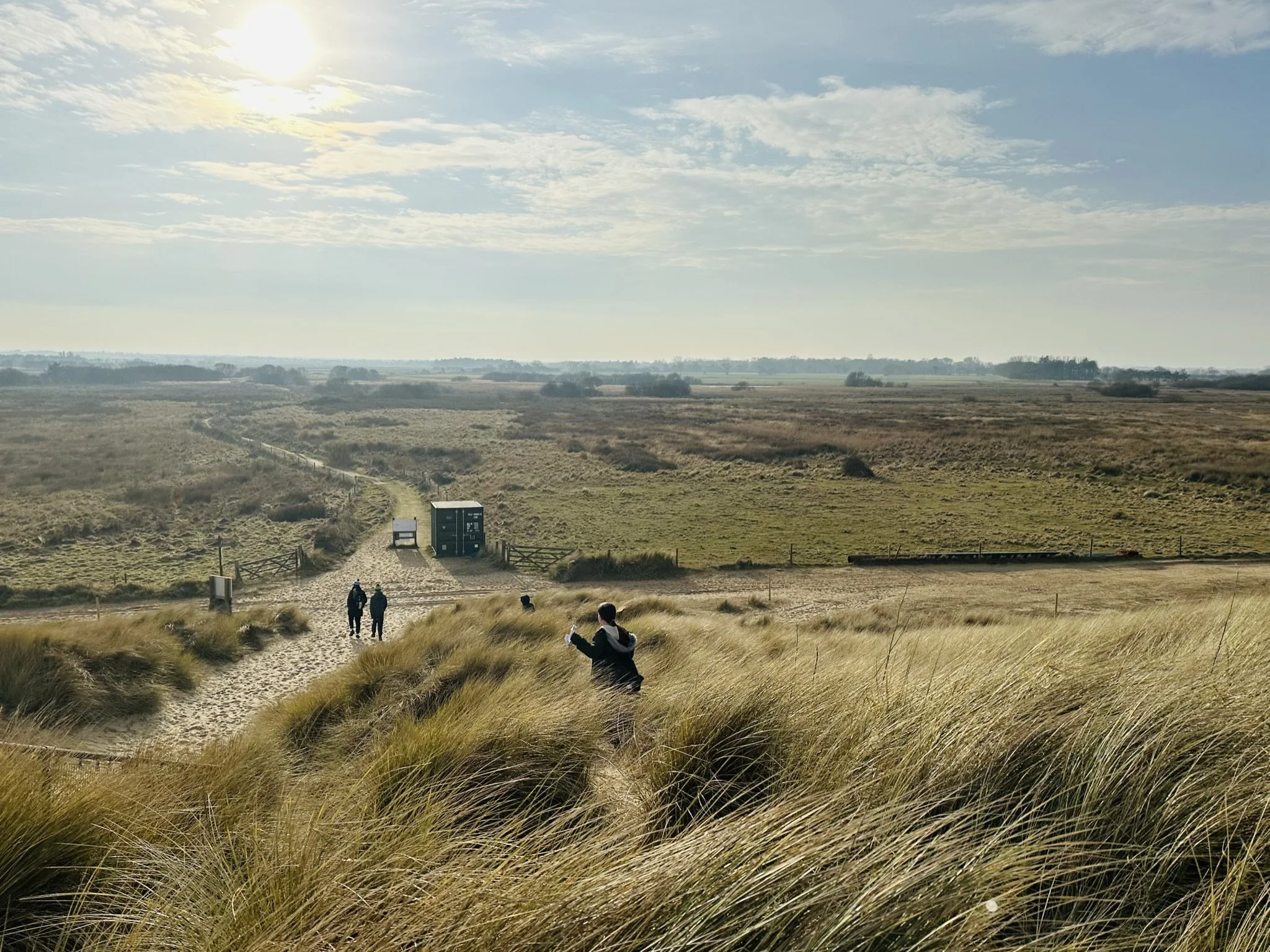 Horsey beach sand dunes and grass in the foreground with four people walking on a sandy trail toward a field, a small black structure, and a distant horizon under a partly cloudy sky.
