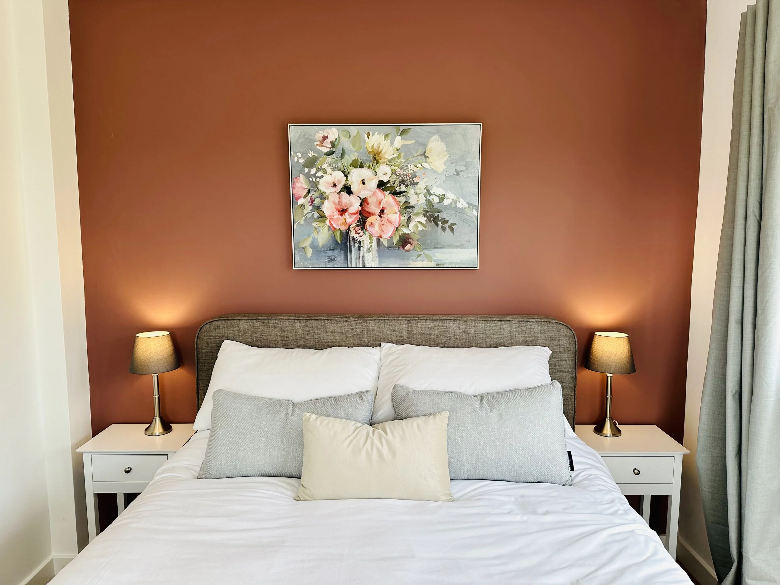 A bedroom with a kingsize bed against a rust-colored accent wall, decorated with a floral painting, two bedside tables with lamps, and five pillows on the bed.
