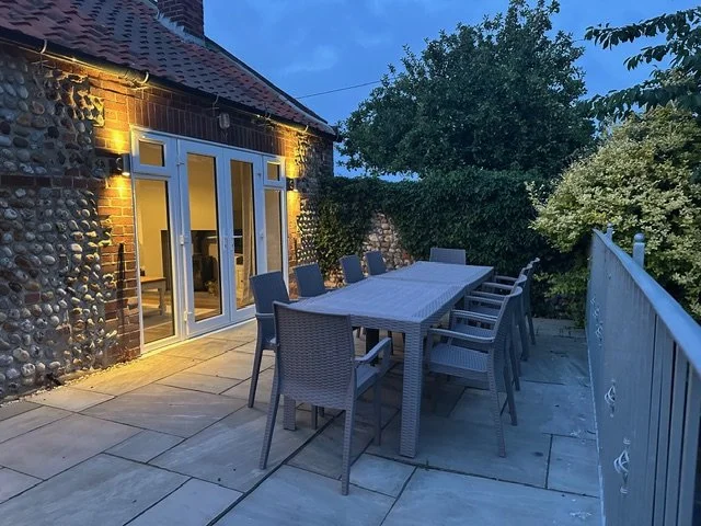 Alfresco dining with the whole family on this beautiful outdoor garden terrace, with a long dining table and several chairs, attached to a brick house with glass doors, surrounded by greenery and illuminated by warm lighting at dusk.