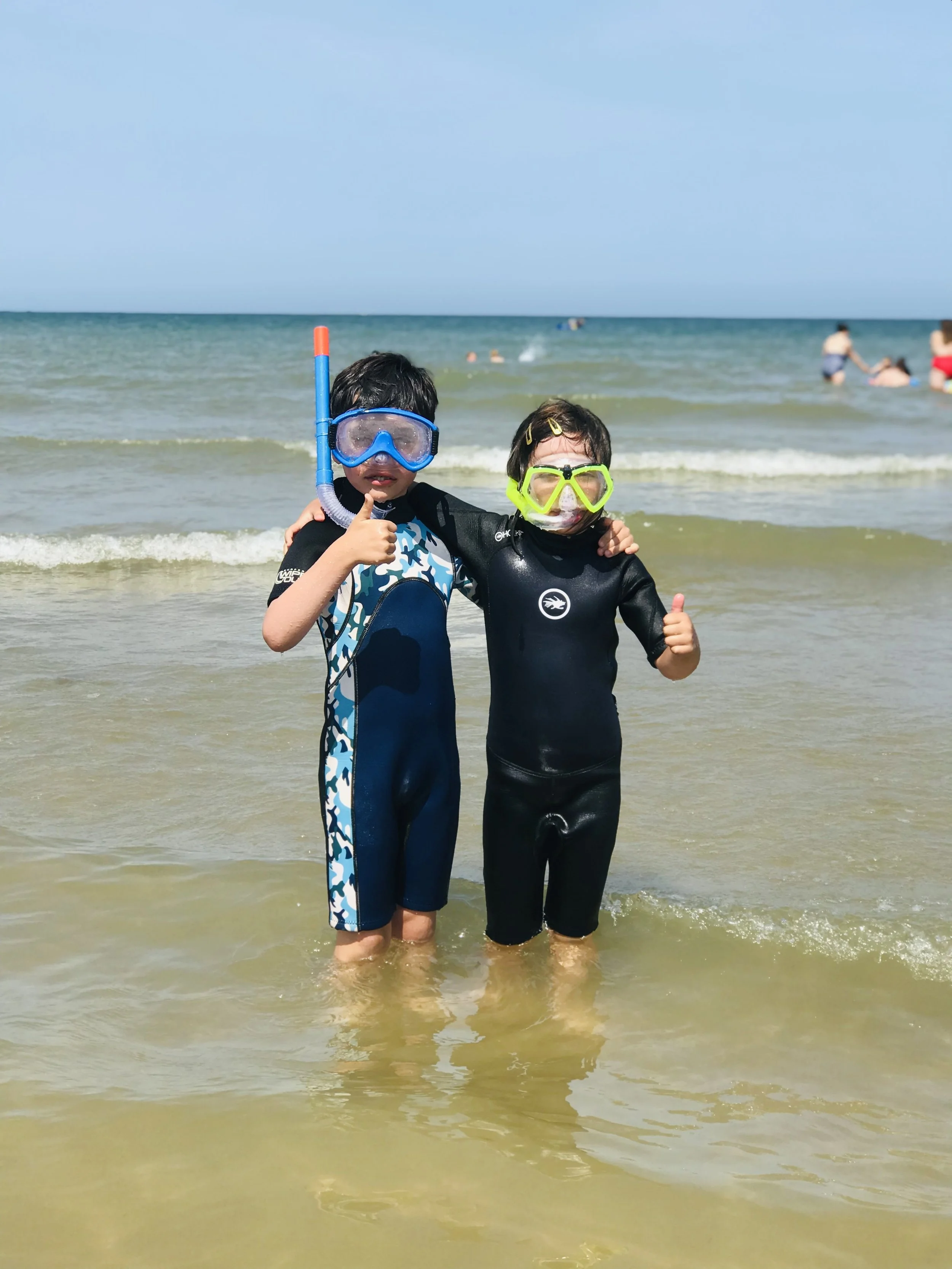 Two children in wetsuits and snorkel gear standing in shallow ocean water on Cromer Beach, by Cromer Pier, giving thumbs up. In the background, other people swimming and playing in the North Sea