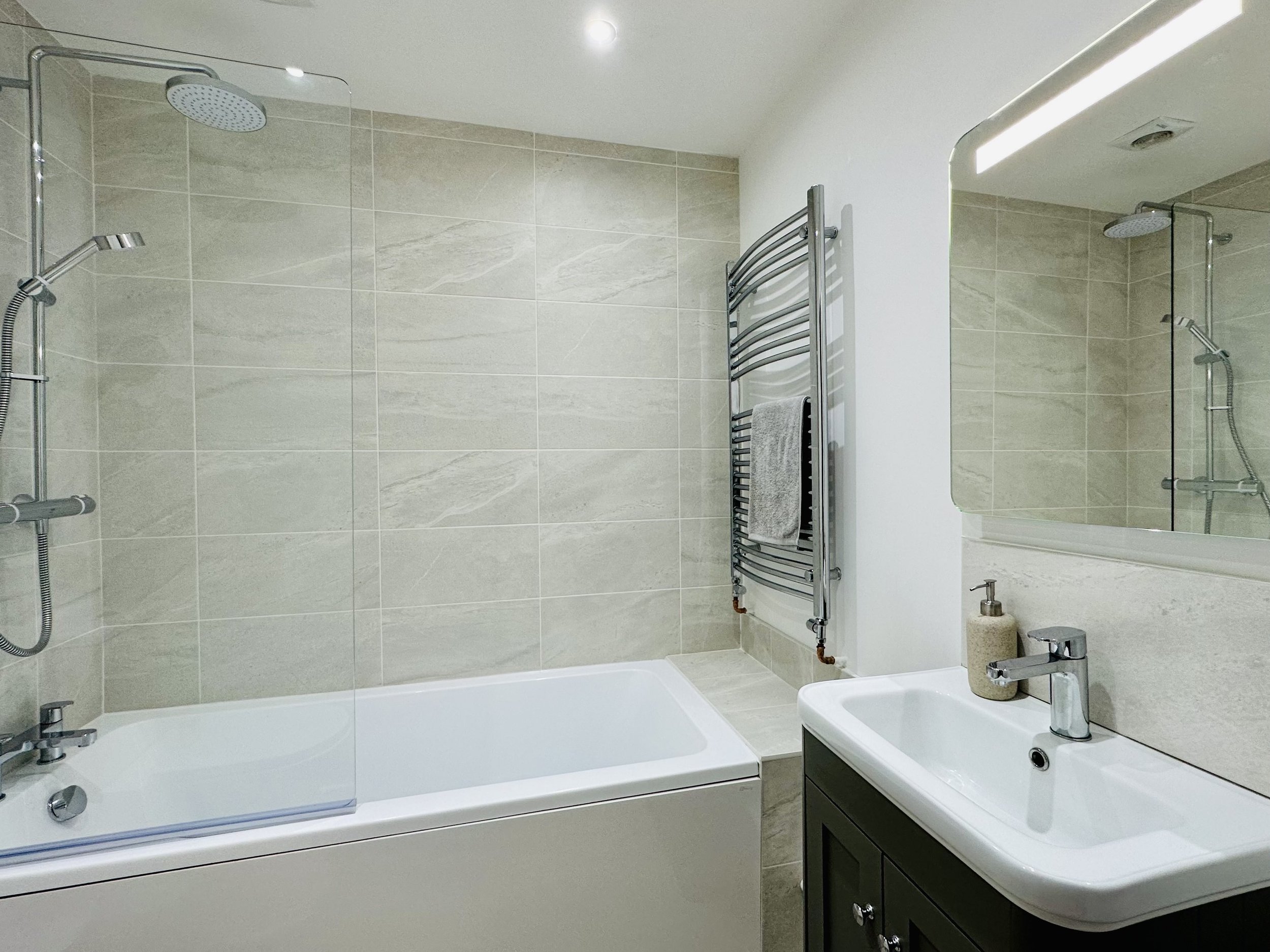 Modern bathroom with beige tiled walls, a white bathtub to wash the kids down after a day at the beach, with a towel warmer, a white sink with a silver tap, a soap dispenser, and a large mirror.