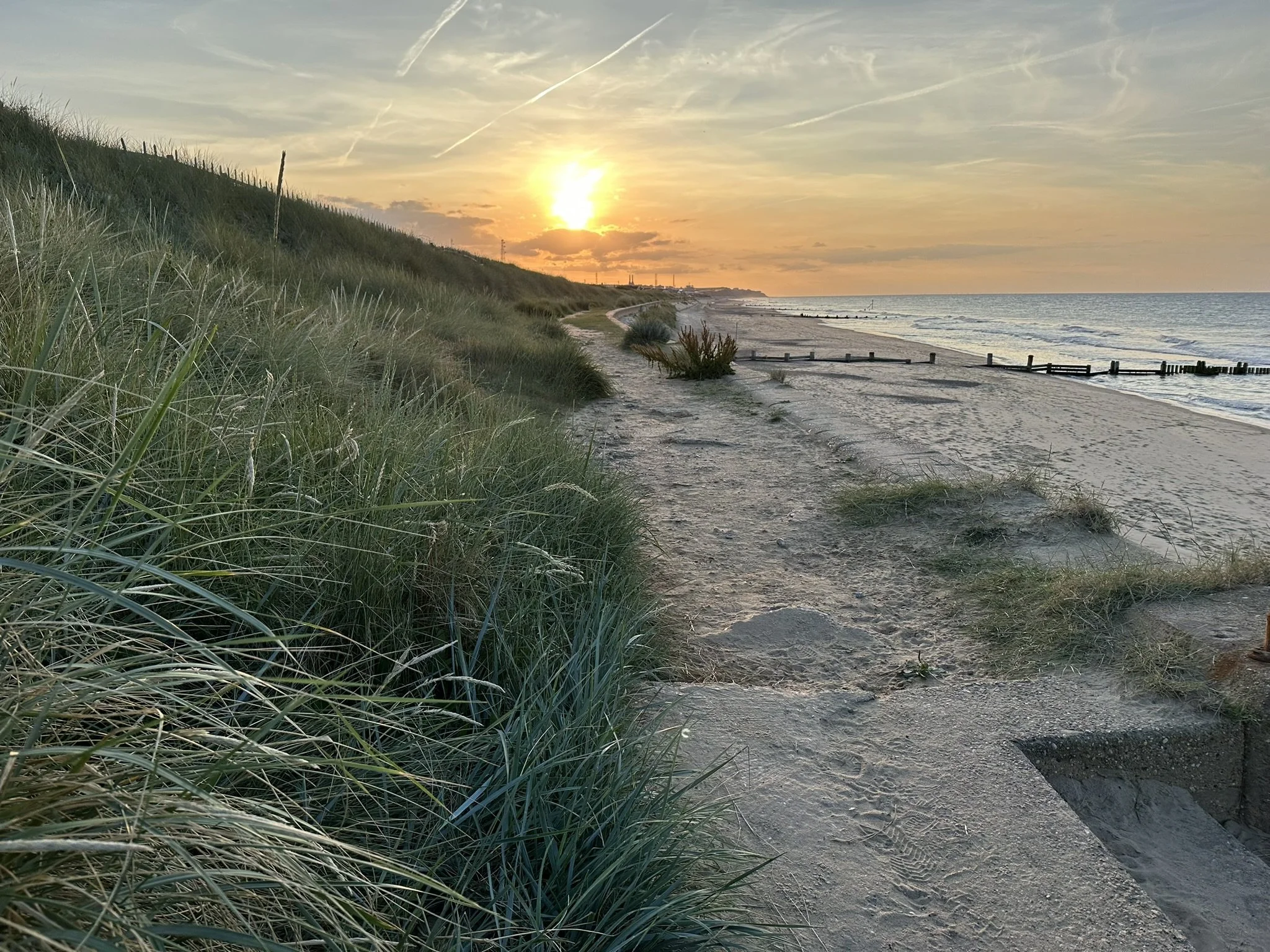 Sunset at Bacton beach (North Norfolk) with grassy dunes on the left and a sandy shoreline on the right, with the ocean and a partly cloudy sky in the background.
