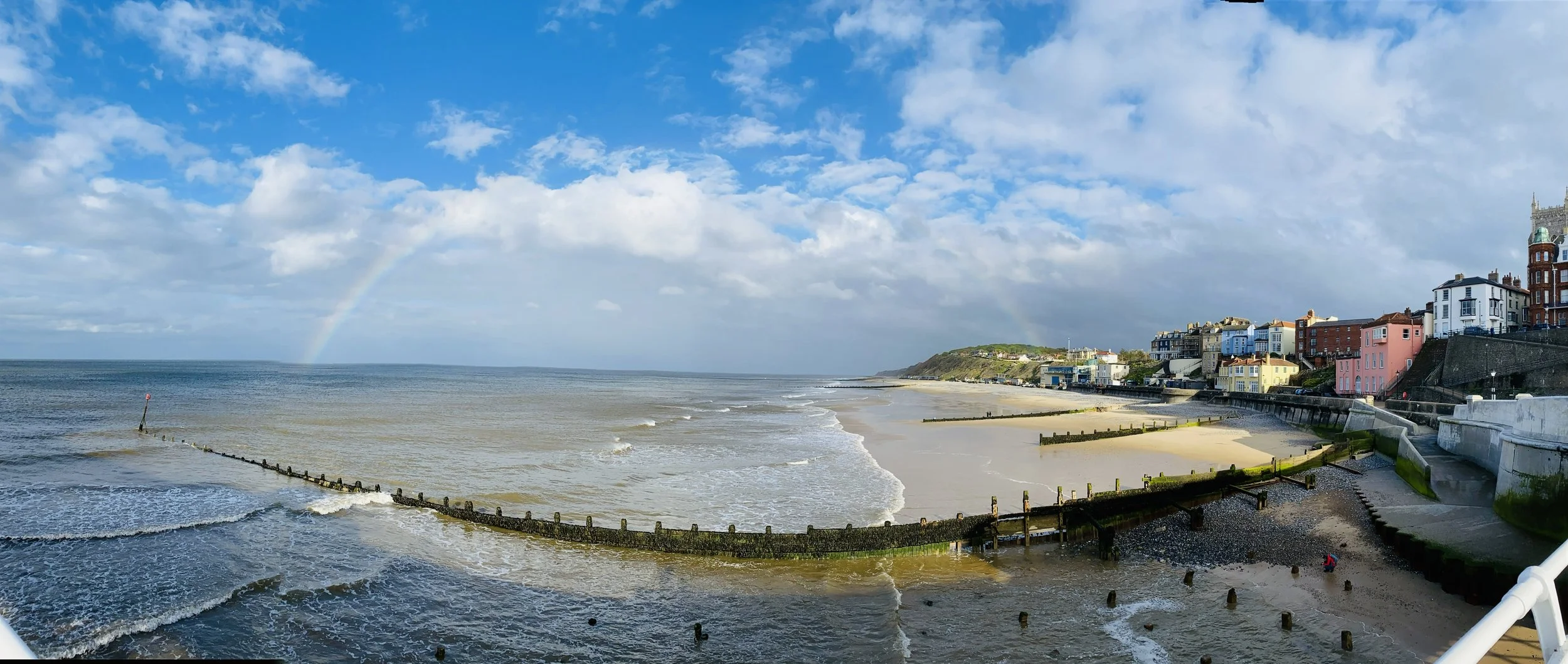 Scenic view of a sandy beach from Cromer Pier (North Norfolk) with gentle waves, colourful buildings lining the coast, cloudy sky with a faint rainbow, and a partly visible person on the beach.