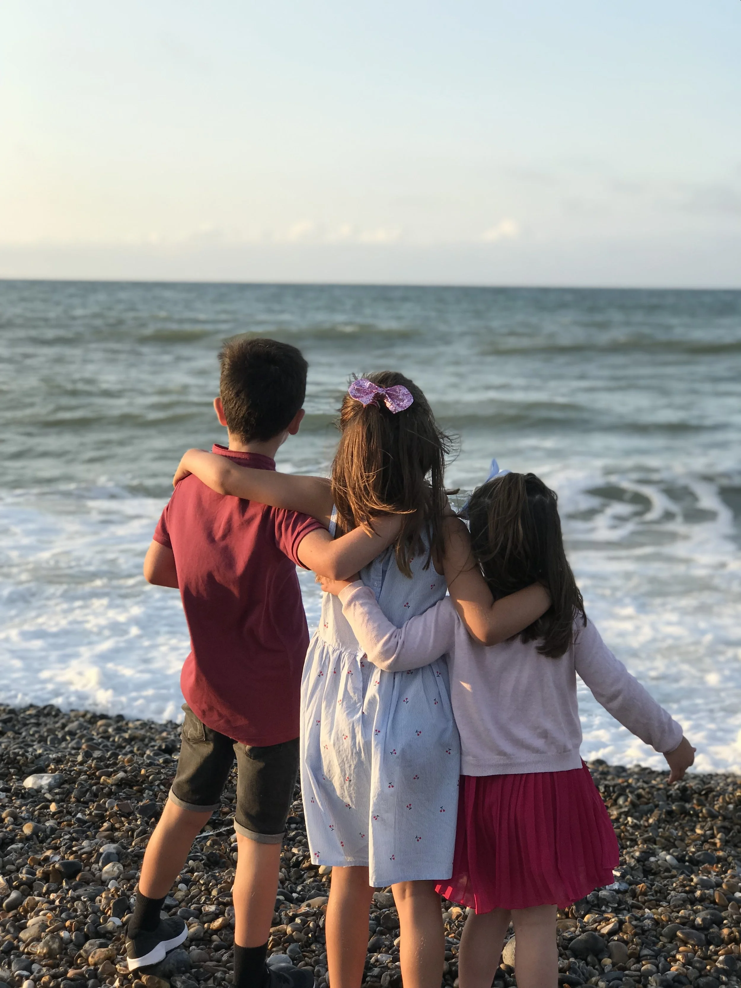 Four children standing on a pebble beach, facing the ocean, embracing each other.