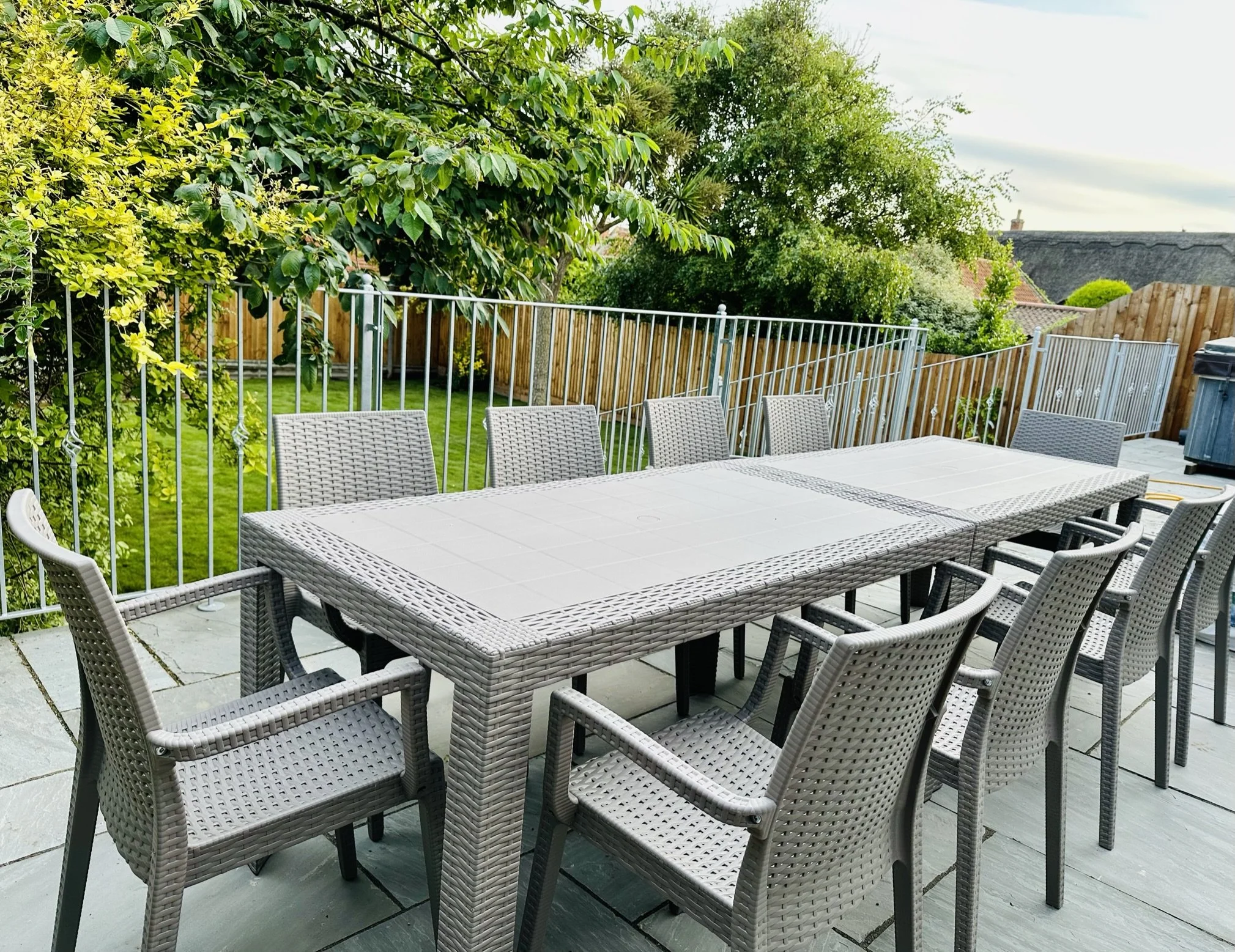 Outdoor patio area for the whole family to eat breakfast outside on holiday, with a large rectangular table and ten woven chairs, surrounded by a metal fence, green trees, and a wooden fence in the background.