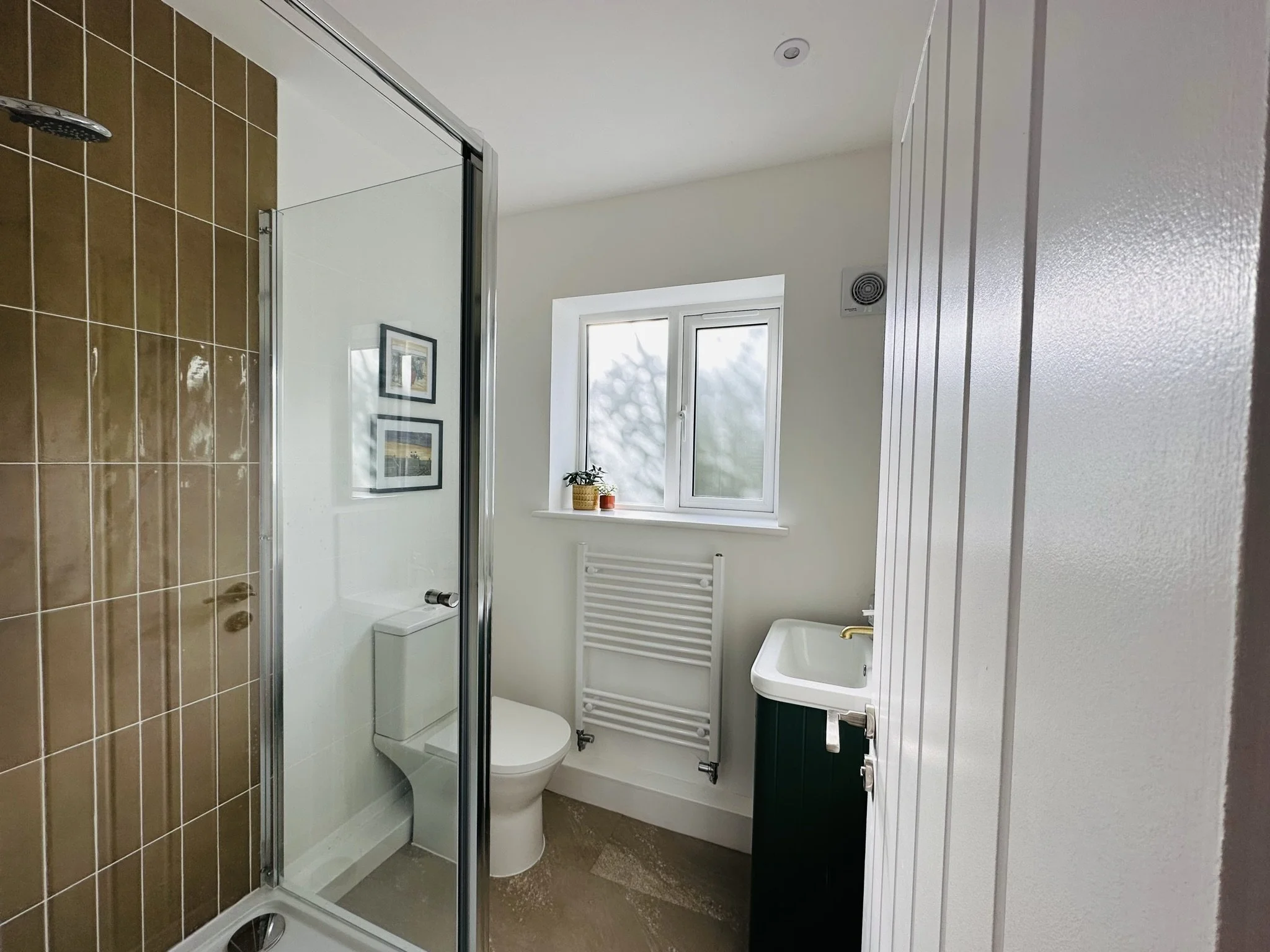 Mustard yellow tiled shower room with green sink cabinet with gold tap, with a glass-enclosed shower on the left, a toilet beside the window, a radiator below the window, and framed pictures on the wall. 