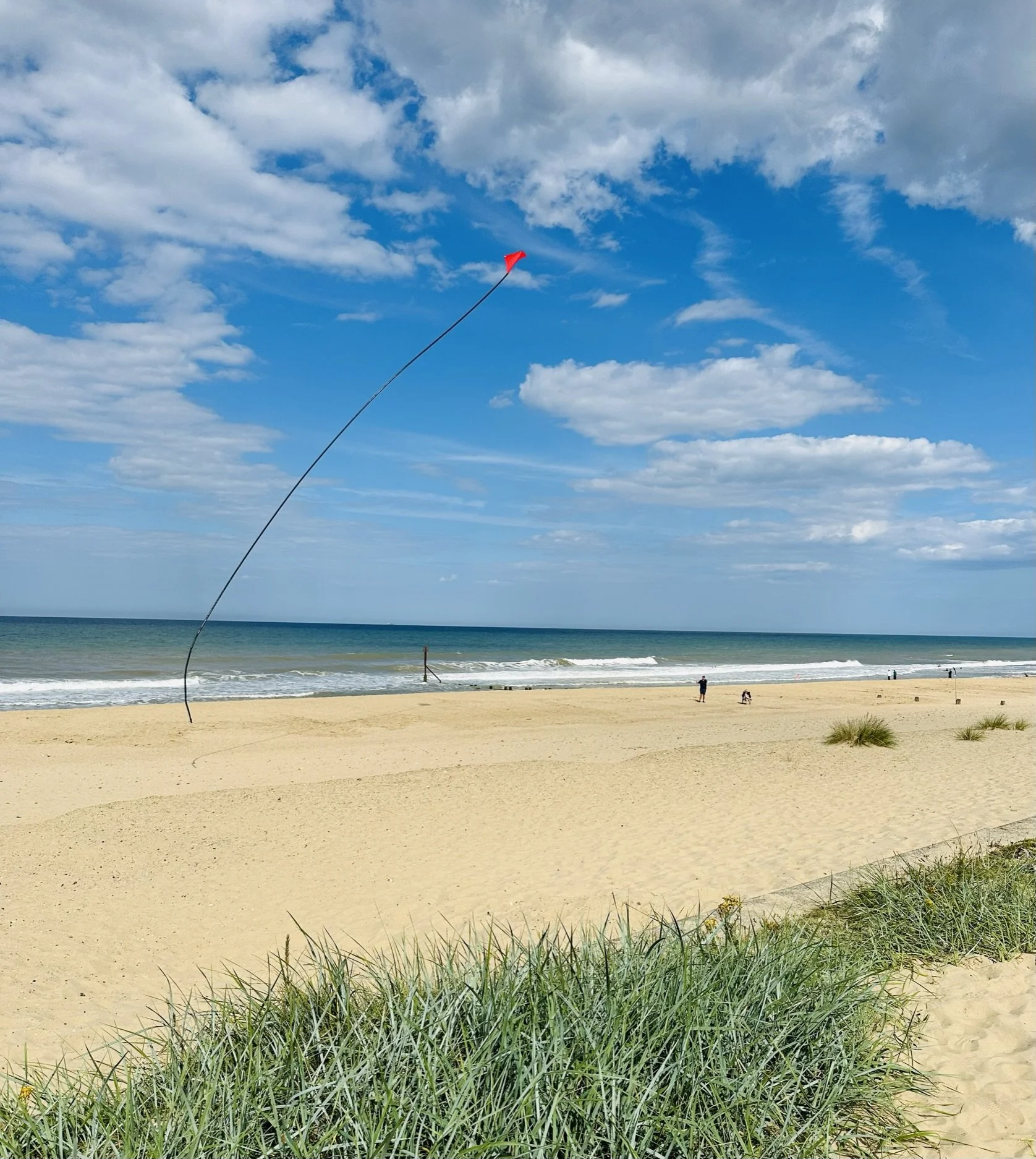 Bacton's sandy beach with some green grass in the foreground, a calm ocean with small waves, a partly cloudy blue sky, and a person flying a red kite.