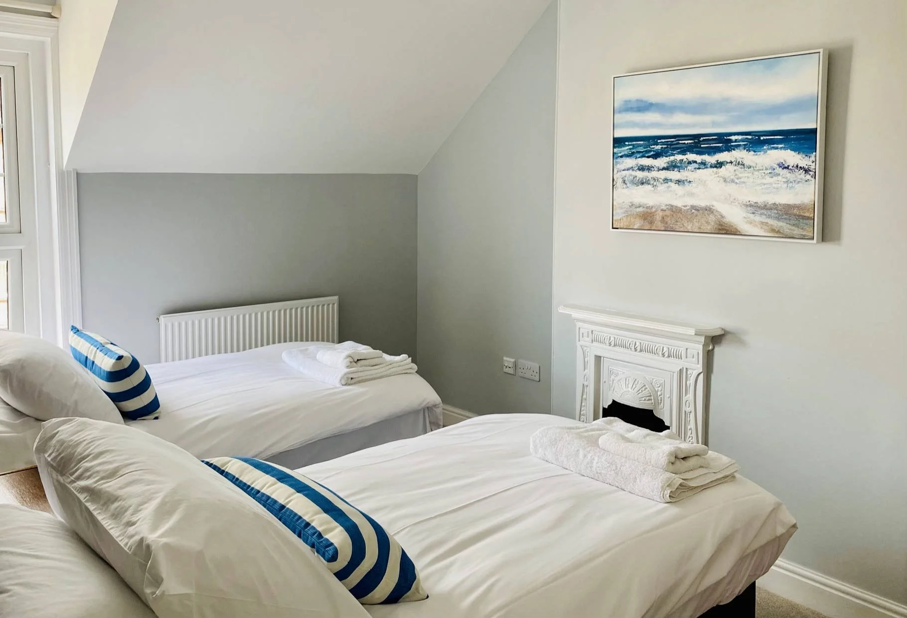 Twin bedroom in Coast House, Cromer, each with white linens, a striped blue and white pillow, and folded towels at the foot. The room has a white decorative fireplace, light colored walls, 