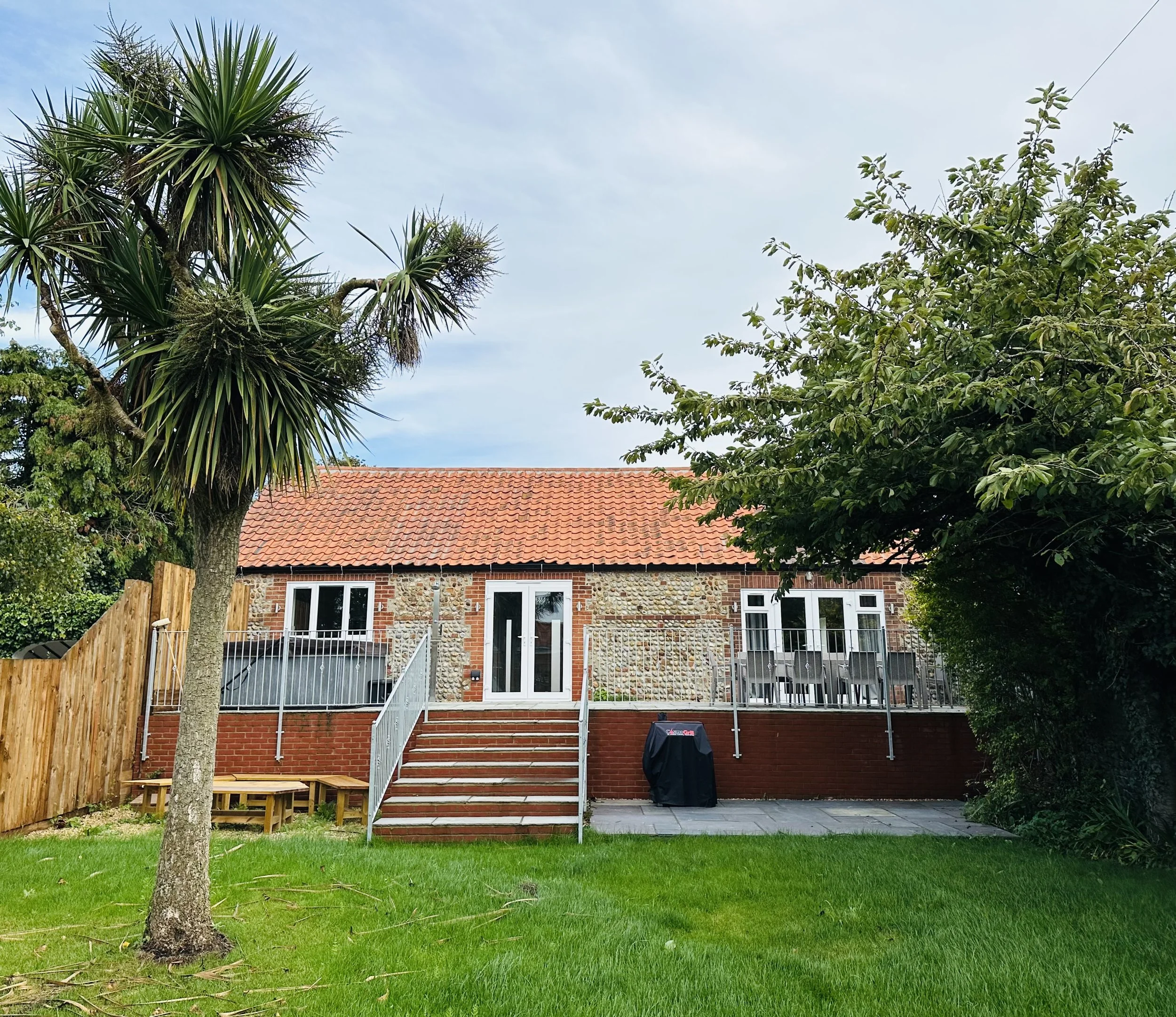 Large lawn are for the children to play whilst on holiday by the sea, a palm tree on the left. The house has white-framed windows and a white door with stairs leading up to it.