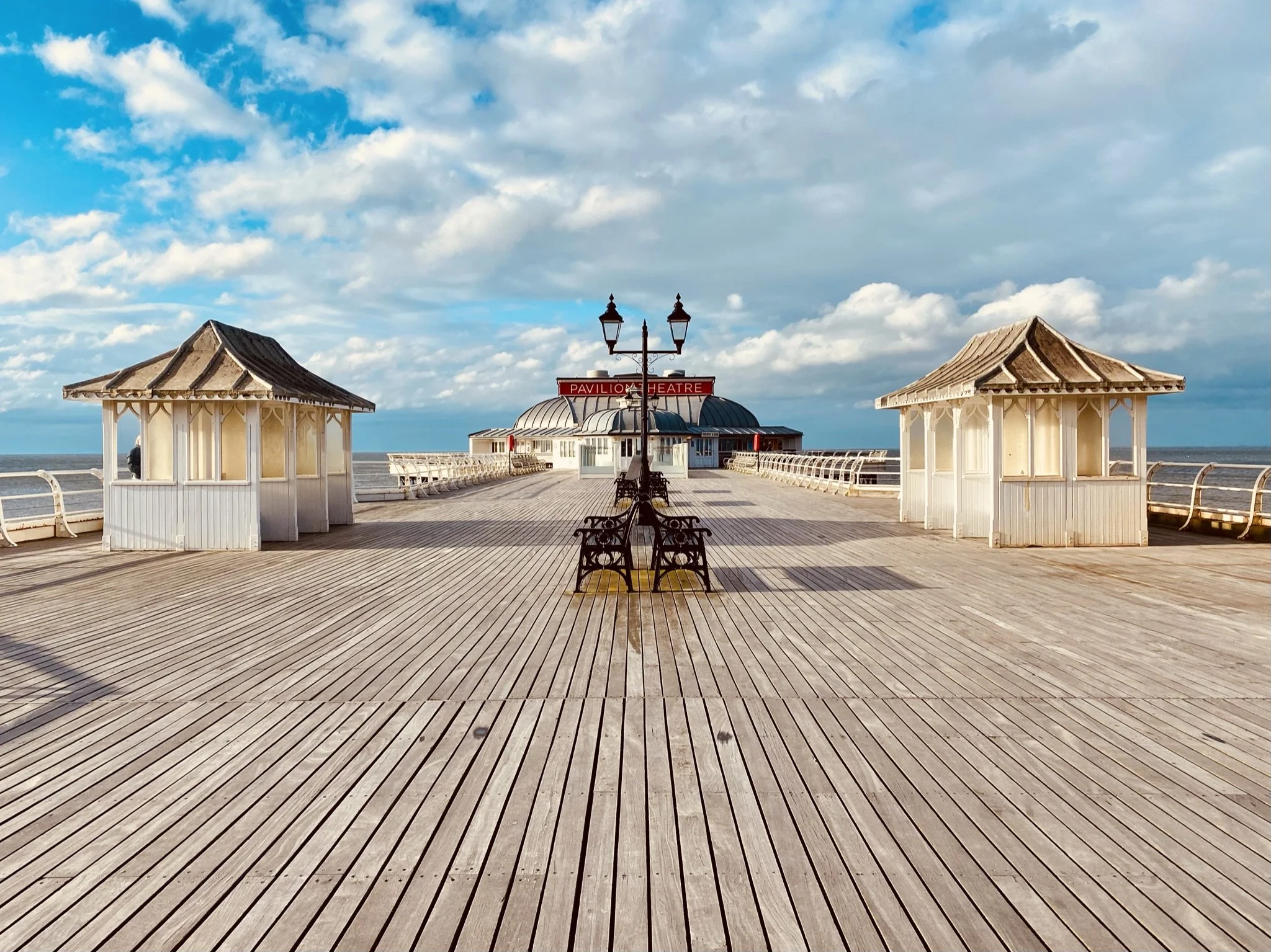 Cromer Pier with wooden floors, two small white pavilions, a lamp post with benches, and a pavilion theater at the end, under a partly cloudy sky, with beautiful views across the North Sea