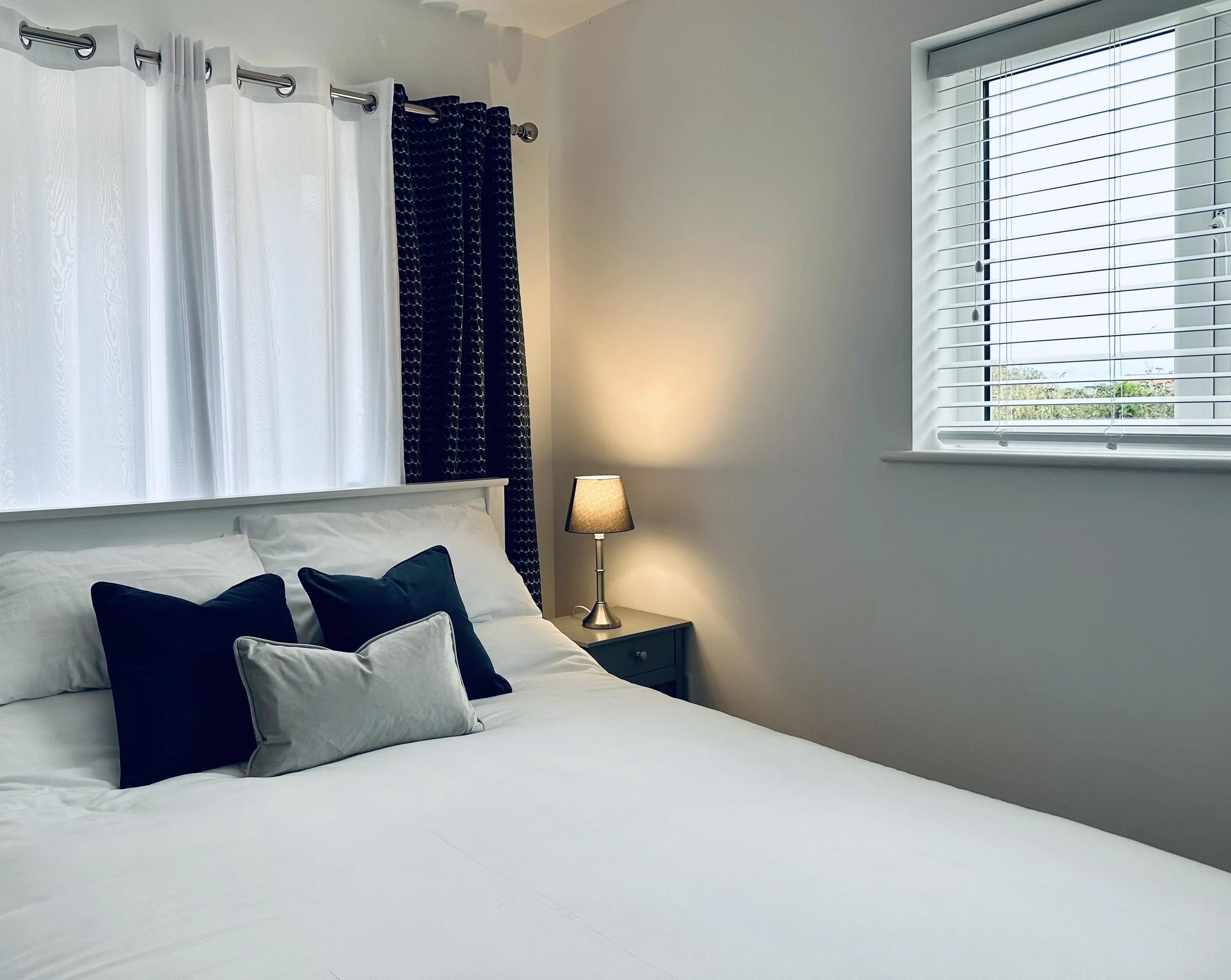 Kingsize bed in white bedroom, decorative pillows, a nightstand with a lamp, and a window with white blinds in Bromholme Cottage, Bacton