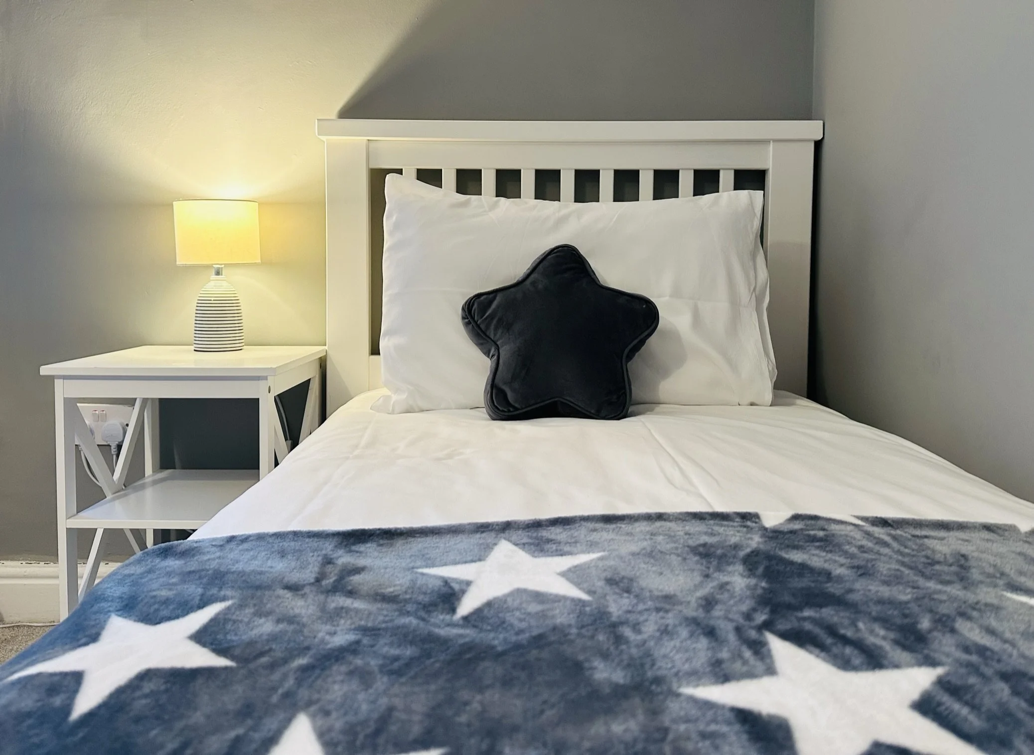 A neatly made bed with white sheets, a black star-shaped pillow, and a star-patterned blanket in navy with white stars. There is a white wooden headboard and a bedside table with a lamp. The wall behind is painted gray.