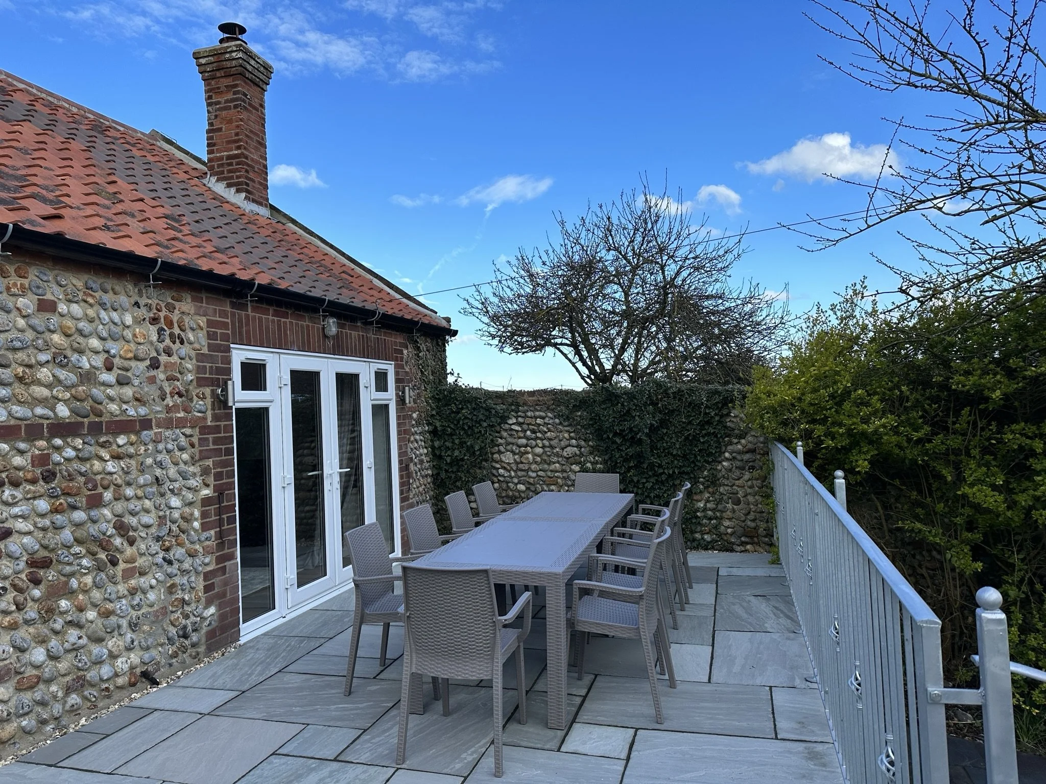 An outdoor patio area with a large dining table and chairs, with space for the whole family, up to 10 people, adjacent to a house with a stone and brick exterior, under a blue sky with some clouds and trees in the background.