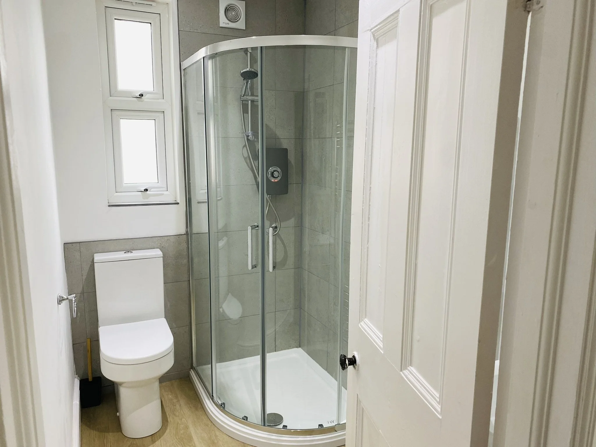 Spacious shower room with a toilet and a glass corner shower enclosure. Two small, frosted windows are above the toilet, and a door is partially open in the foreground.