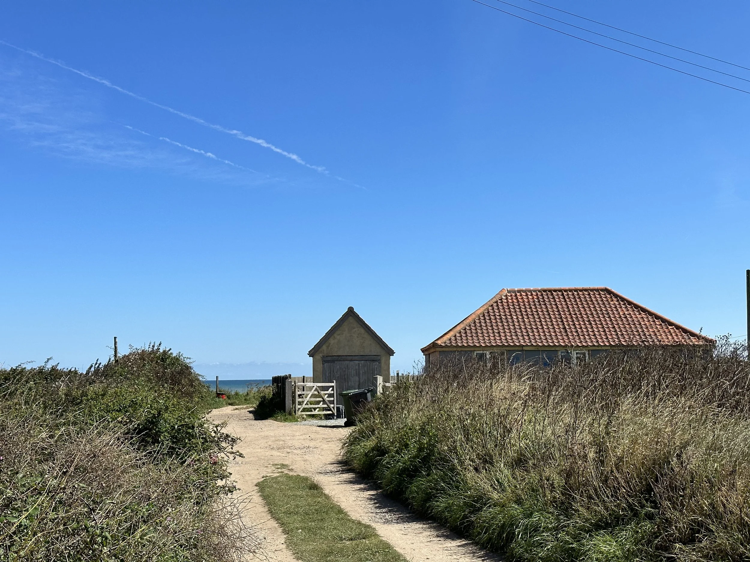 A sandy pathway leading to Bacton's sandy beach, two small beach houses under a bright blue sky with some clouds and contrails.