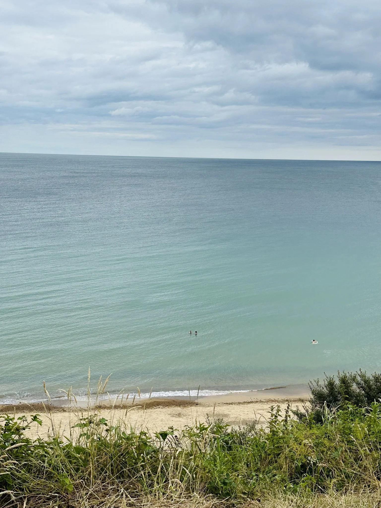 Overstrand beach, North Norfolk, a beach scene with greenish-blue ocean water, sandy shore, and cloudy sky, with two people swimming and one person on a float in the water.