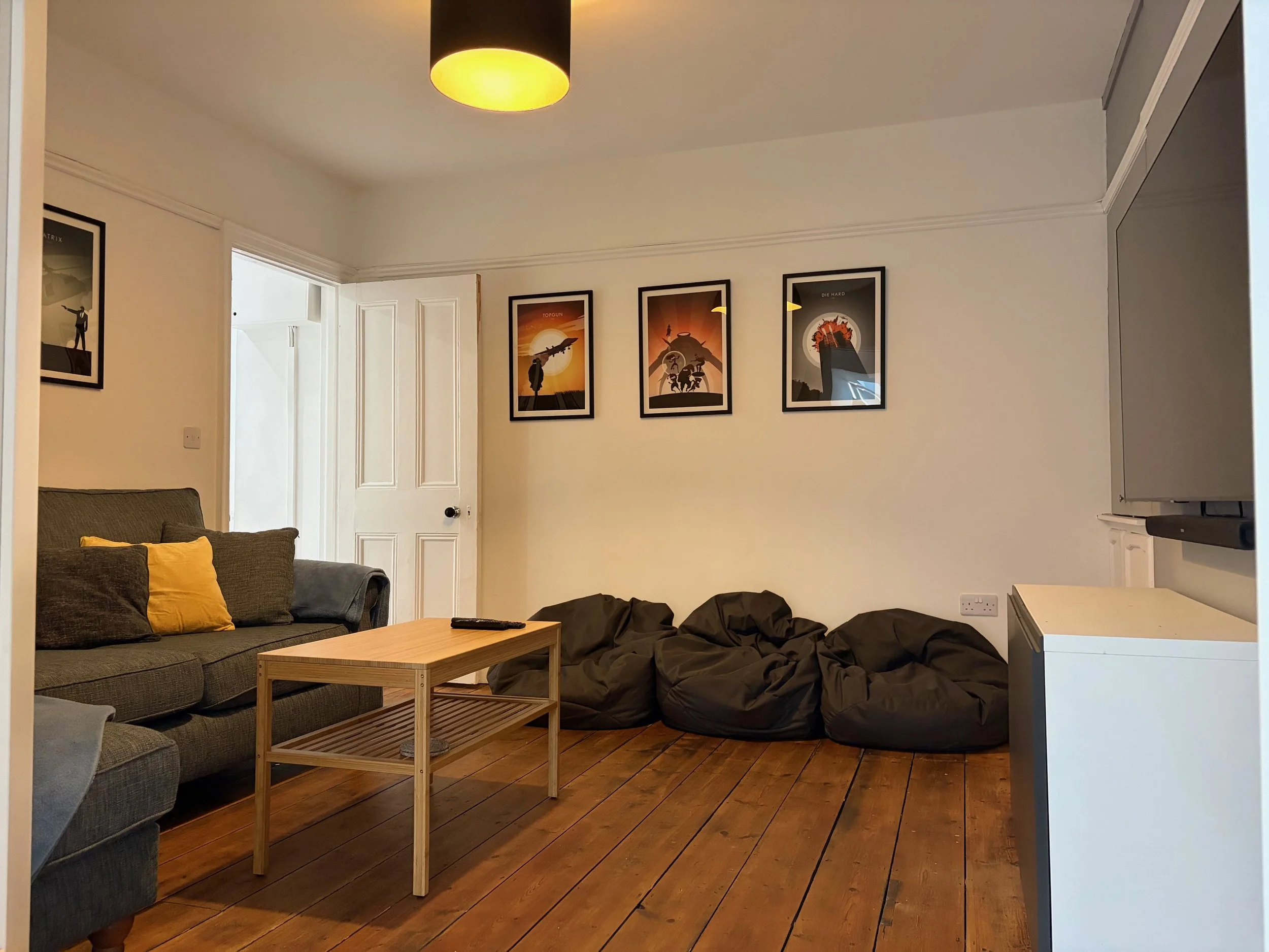 Cinema room in Coast House, Cromer, with a gray sofa with yellow and gray pillows, a wooden coffee table with a remote control, three black bean bags on the wooden floor, framed artwork on the wall, and a partially open door.