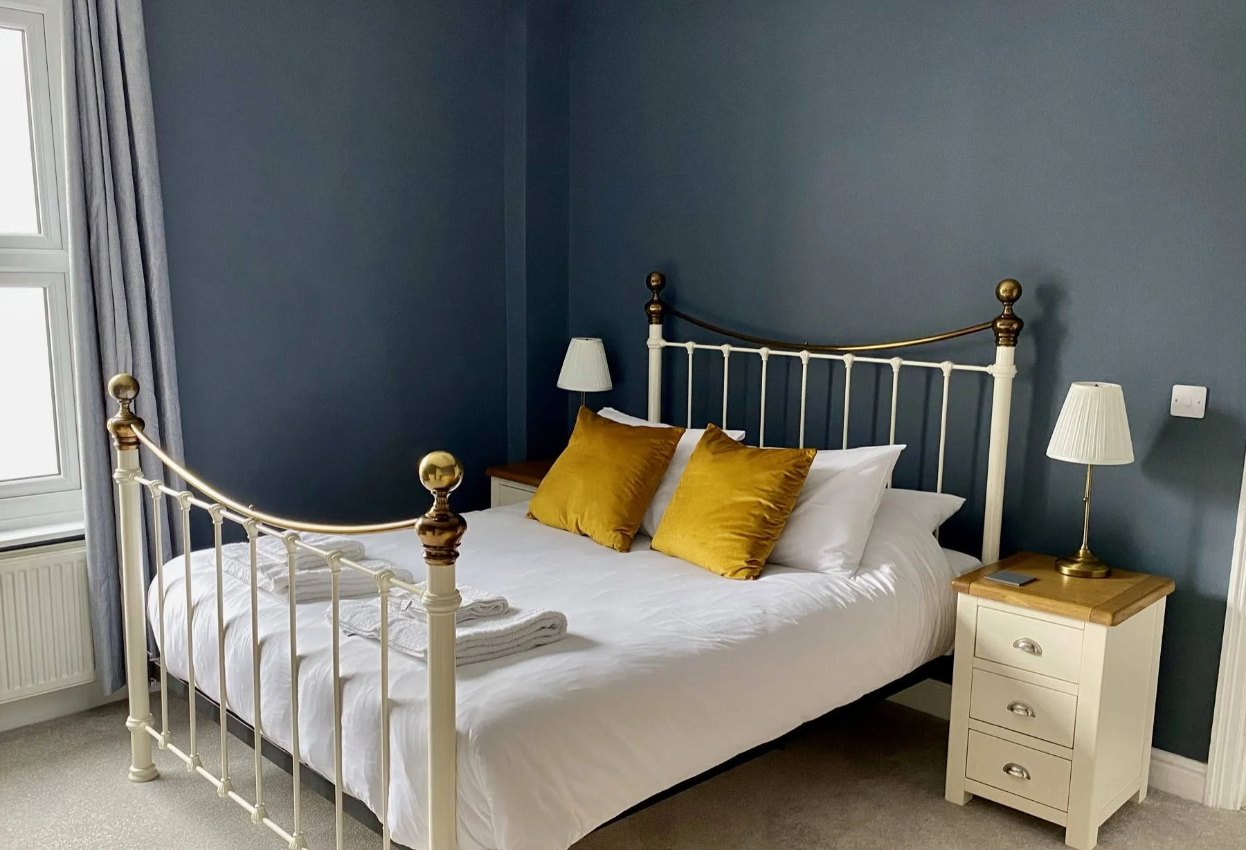 Large bedroom in Coast House, Cromer, with a white metal bed frame kingsize bed, blue walls, a wooden nightstand with a brass lamp, and pillows including two yellow ones, near a window with curtains.