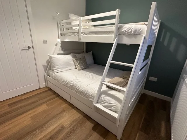 A good bedroom for the children to stay or parents with a child, with white double bunk bed with a lower bed and an upper bed accessed by a ladder, in a room with hardwood floors and a green accent wall.