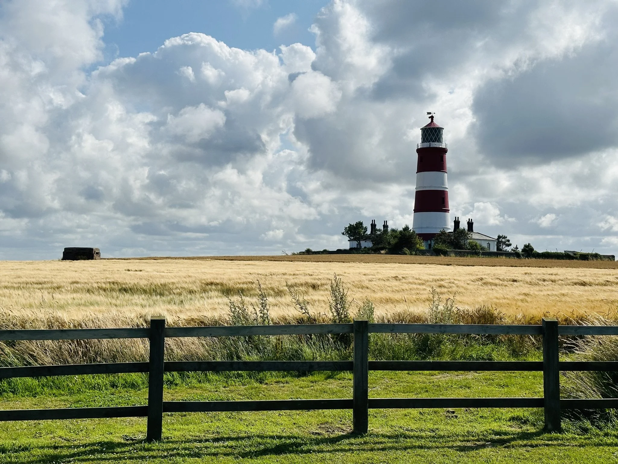 Happisburgh Lighthouse on a grassy hill overlooking a wheat field, with a wooden fence in the foreground and a partly cloudy sky overhead, with play area, toilets and path down to the sandy beach below