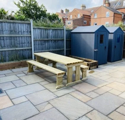 Spacious gardenin Cromer,  area with picnic table, wooden benches on a paved outdoor area, with two blue storage sheds and a wooden fence in the background.