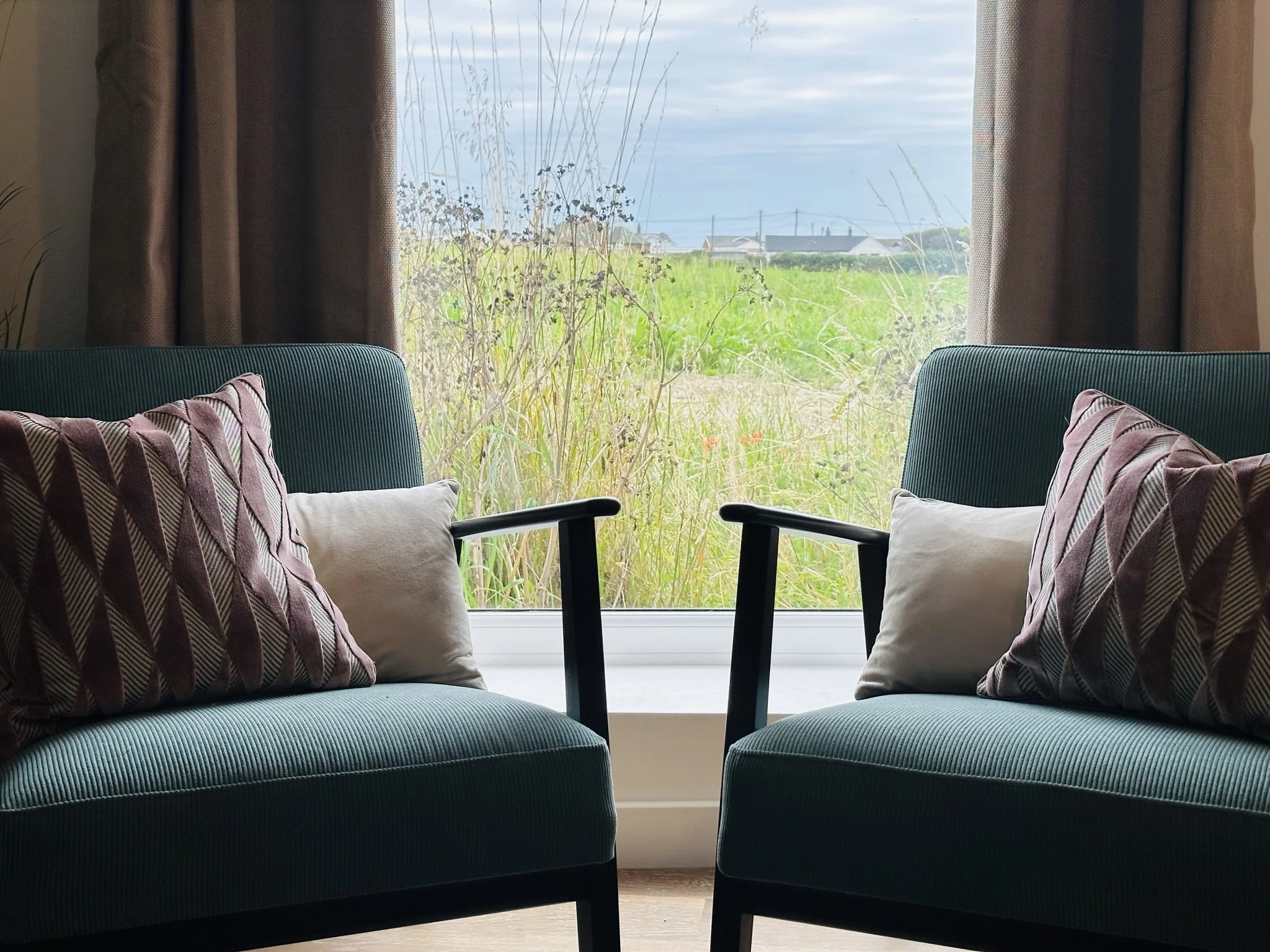 Two teal armchairs overlooking the Barley field, with patterned and plain pillows facing each other in front of a large window with a view of tall grass and a cloudy sky outside.