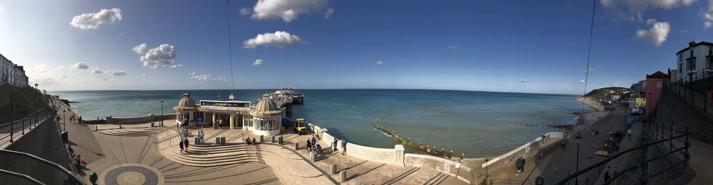 Panoramic view of a seaside town of Cromer with Cromer pier extending into the ocean, a small building with domed roofs at the entrance, and people walking along the promenade on a sunny day with scattered clouds.