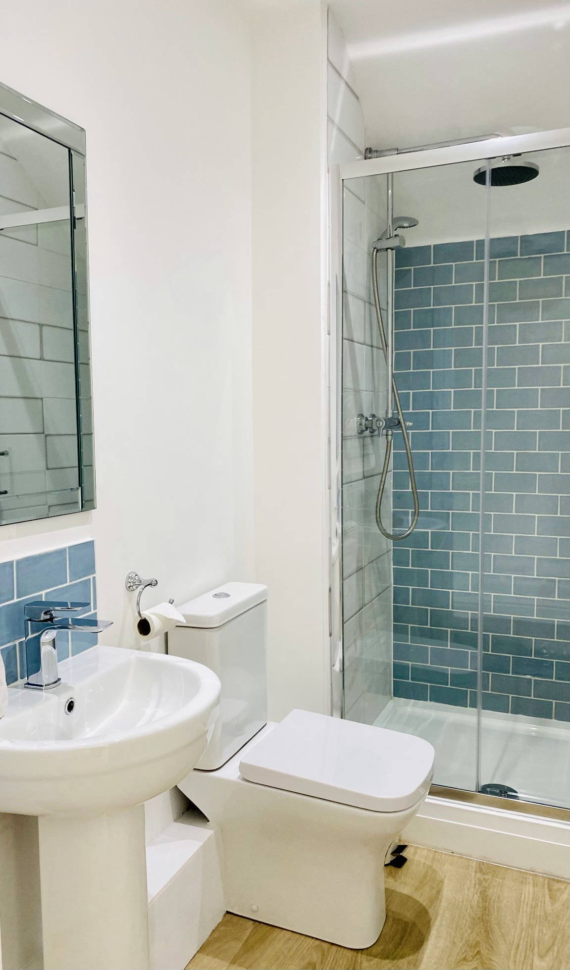 A small modern shower room with a white sink, a toilet, and a glass-enclosed shower with blue subway tiles reminding you of the sea.