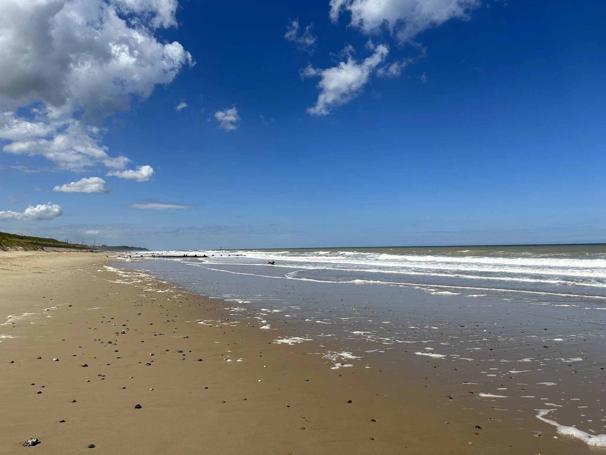 Bacton's sandy beach with small rocks, gentle waves approaching the shore, and a blue sky with scattered clouds.