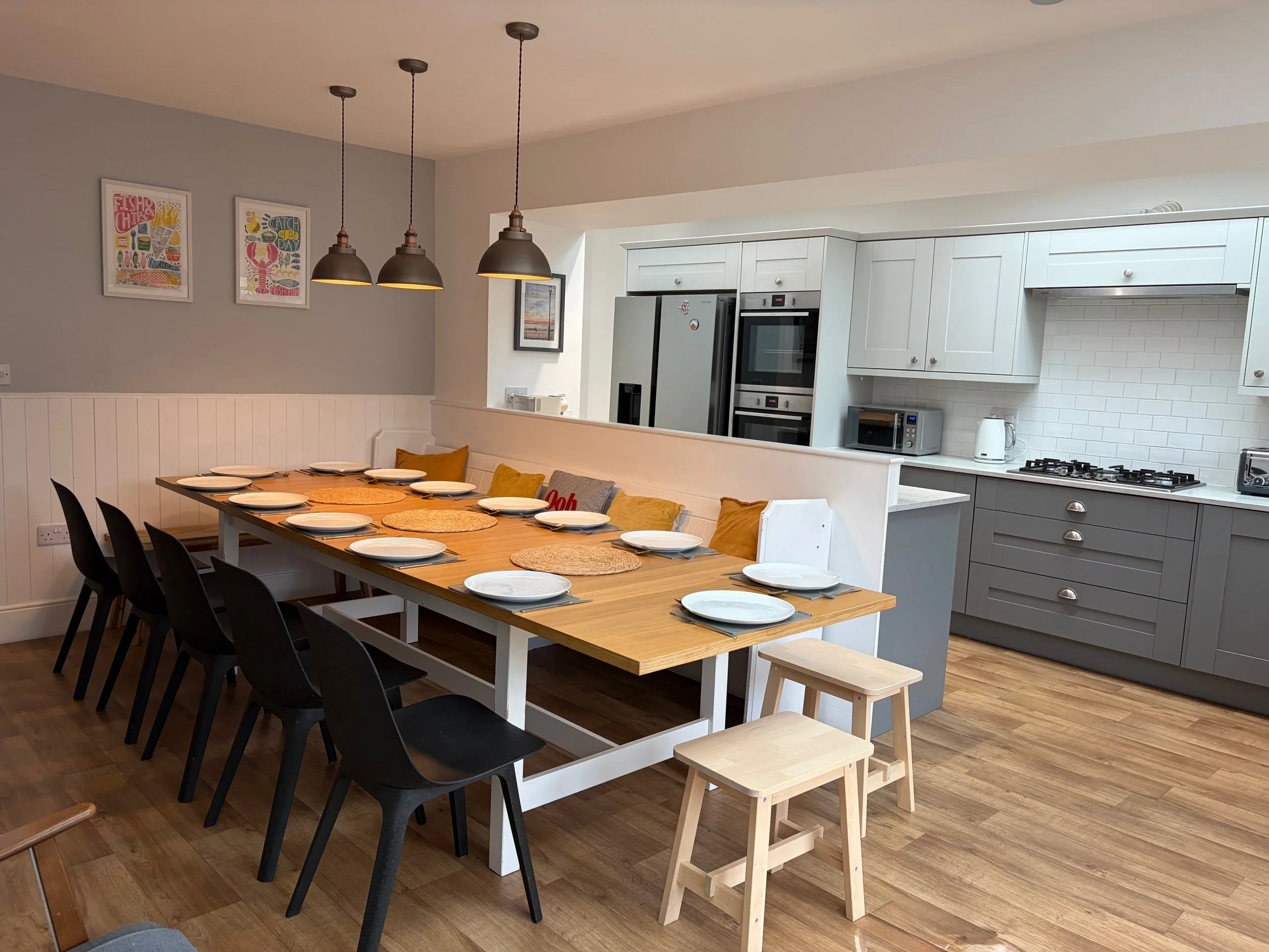A large dining table set with plates, cups, and placemats in a fully equipped modern kitchen with white and gray cabinets, a refrigerator, oven, microwave, and stovetop, with plenty of space for the whole family