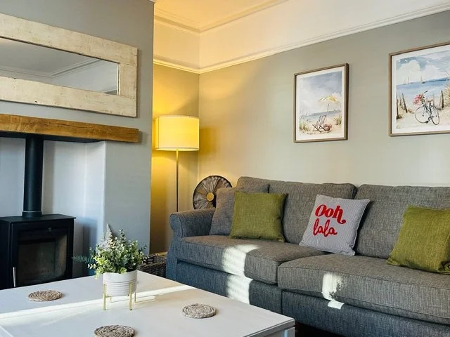 Spacious living room at Coast House, Cromer, with a gray sofa, green and gray pillows, wall art of beach scenes, a yellow floor lamp, a white coffee table with decorative coaster, and a log burner.