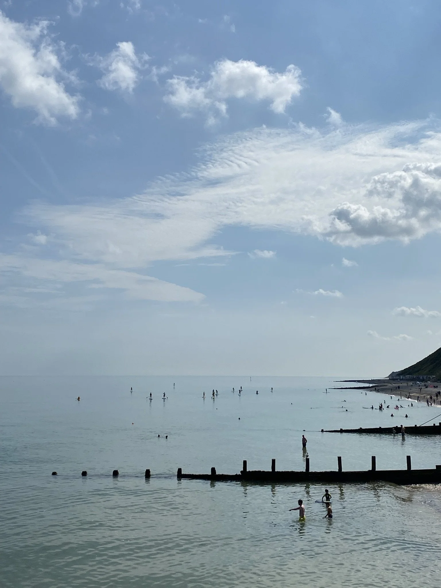 Cromer beach scene in the summer with people swimming and paddle boarding in calm water, a sandy shoreline with some damage, and a hillside in the distance.