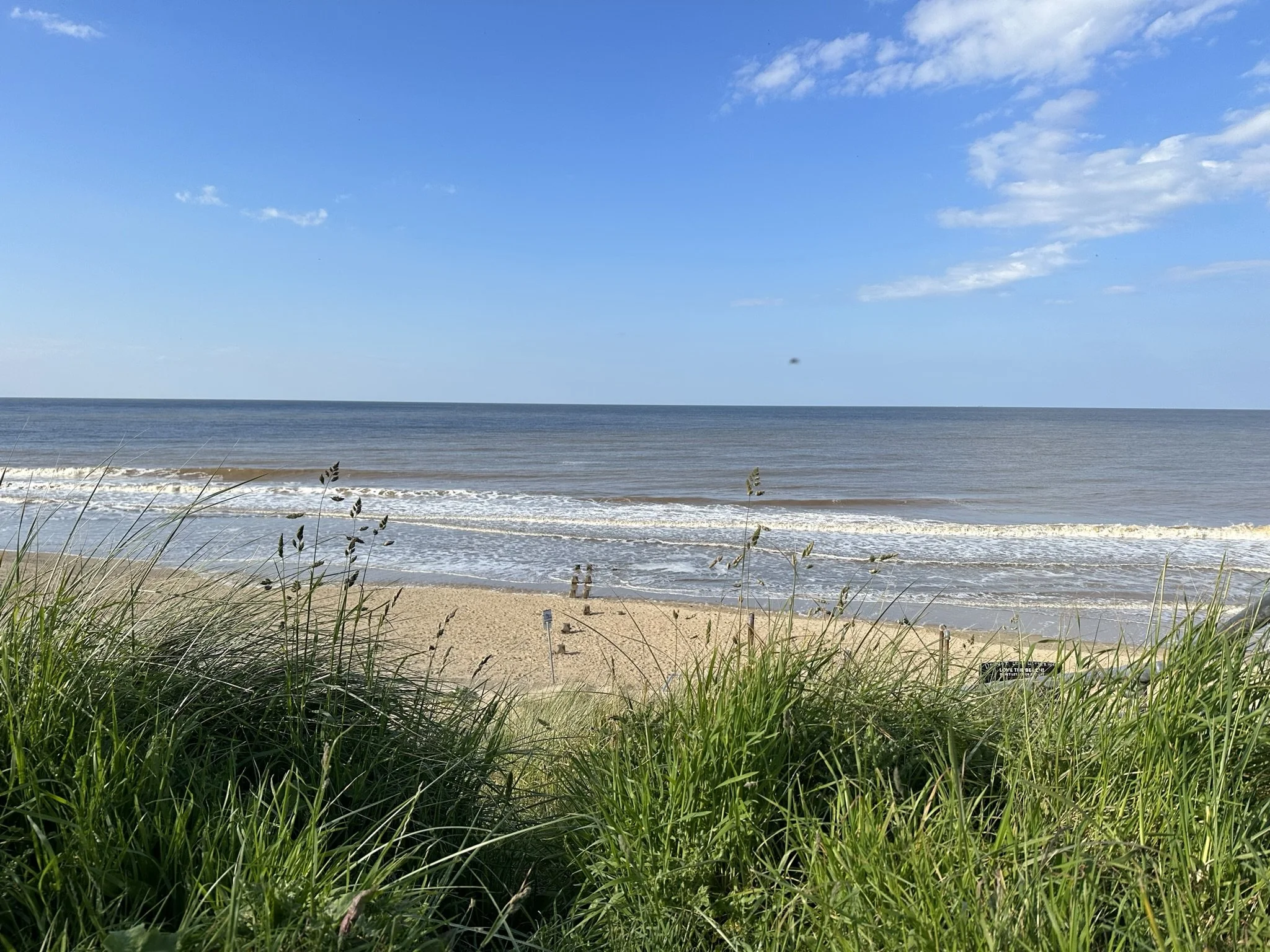 View of Bacton's sandy beach with green grass in the foreground, ocean waves, and a blue sky with clouds.