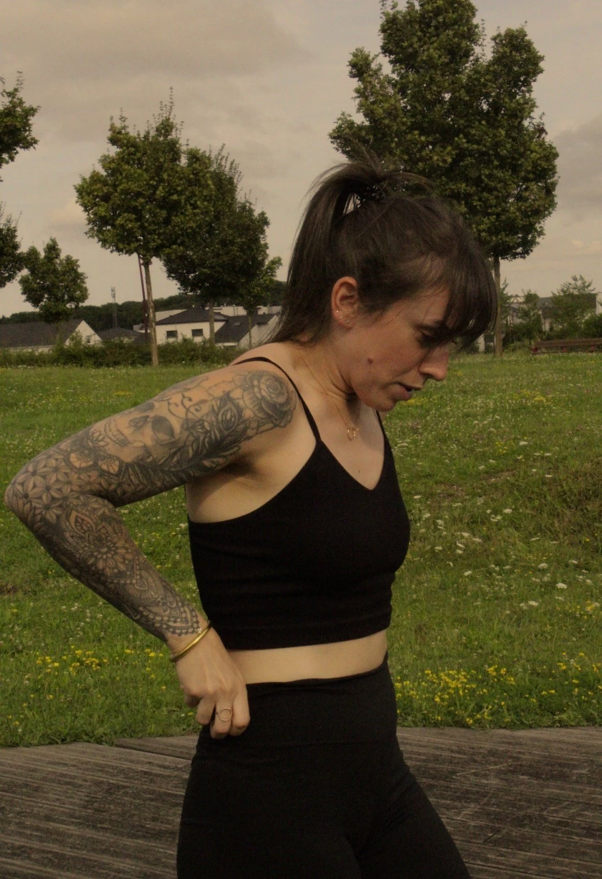 Une femme avec des tatouages et en tenue noire, observant son ventre, dans un parc avec des arbres et une maison en arrière-plan.