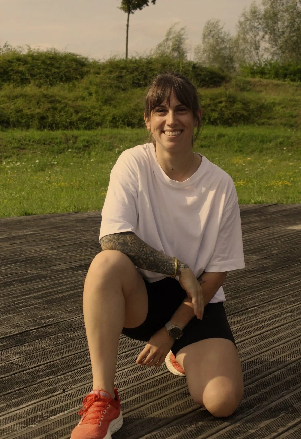 Jeune femme souriante, assise sur un plancher en bois en plein air, portant une t-shirt blanc, un short noir, des baskets rouges, et des accessoires comme une montre et des bracelets, avec un fond de verdure et des arbres.