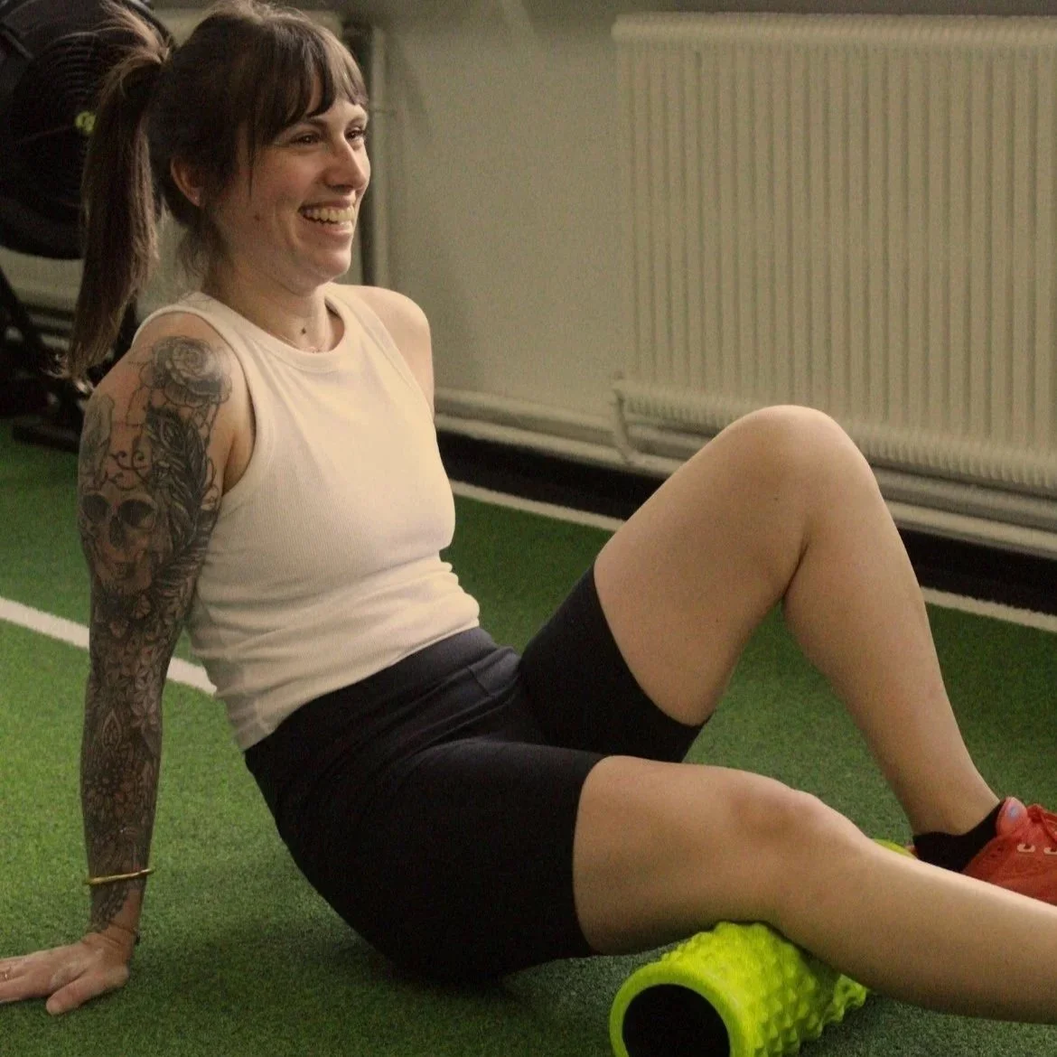 Une femme souriante en tenue de sport utilisant un rouleau de massage en mousse sur une surface en herbe artificielle.