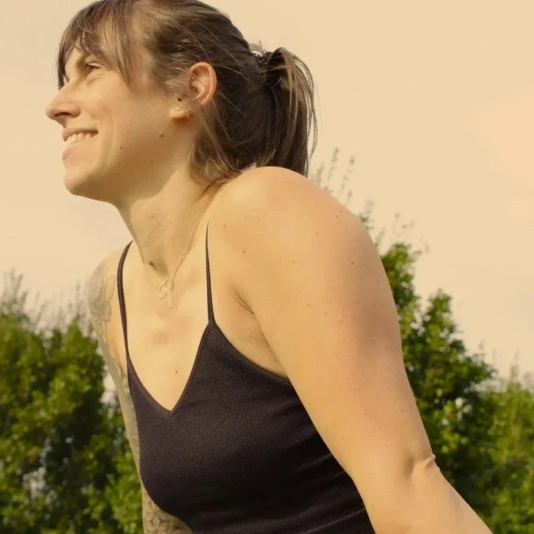 Une femme souriante portant un haut noir à fines bretelles, dehors avec un ciel clair et des arbres en arrière-plan.