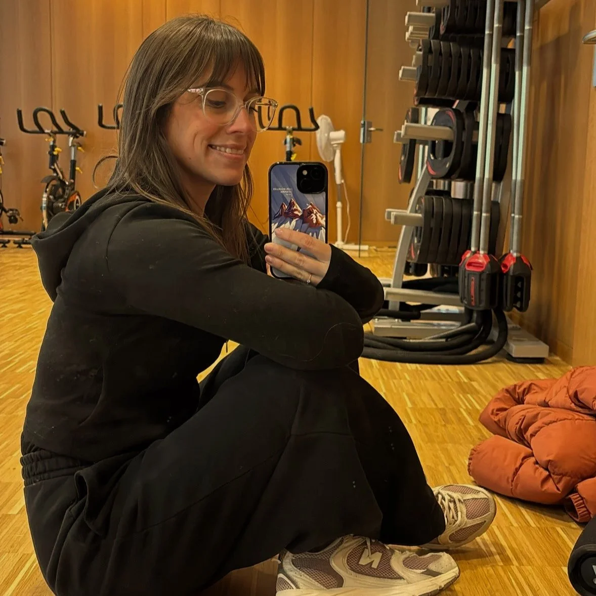 Femme prenant un selfie dans un vestiaire ou une salle de sport avec des équipements d'entraînement derrière elle.