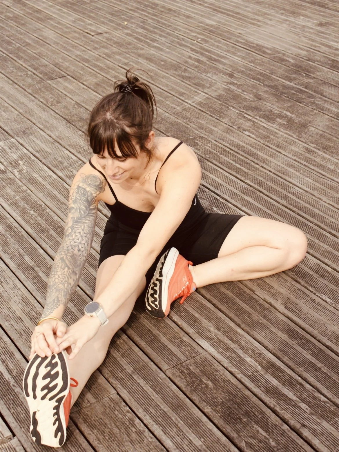 Une femme assise sur un sol en bois, en Yoga, s'étirant avec une jambe tendue, portant une robe noire, sneakers orange, un bracelet et une montre.