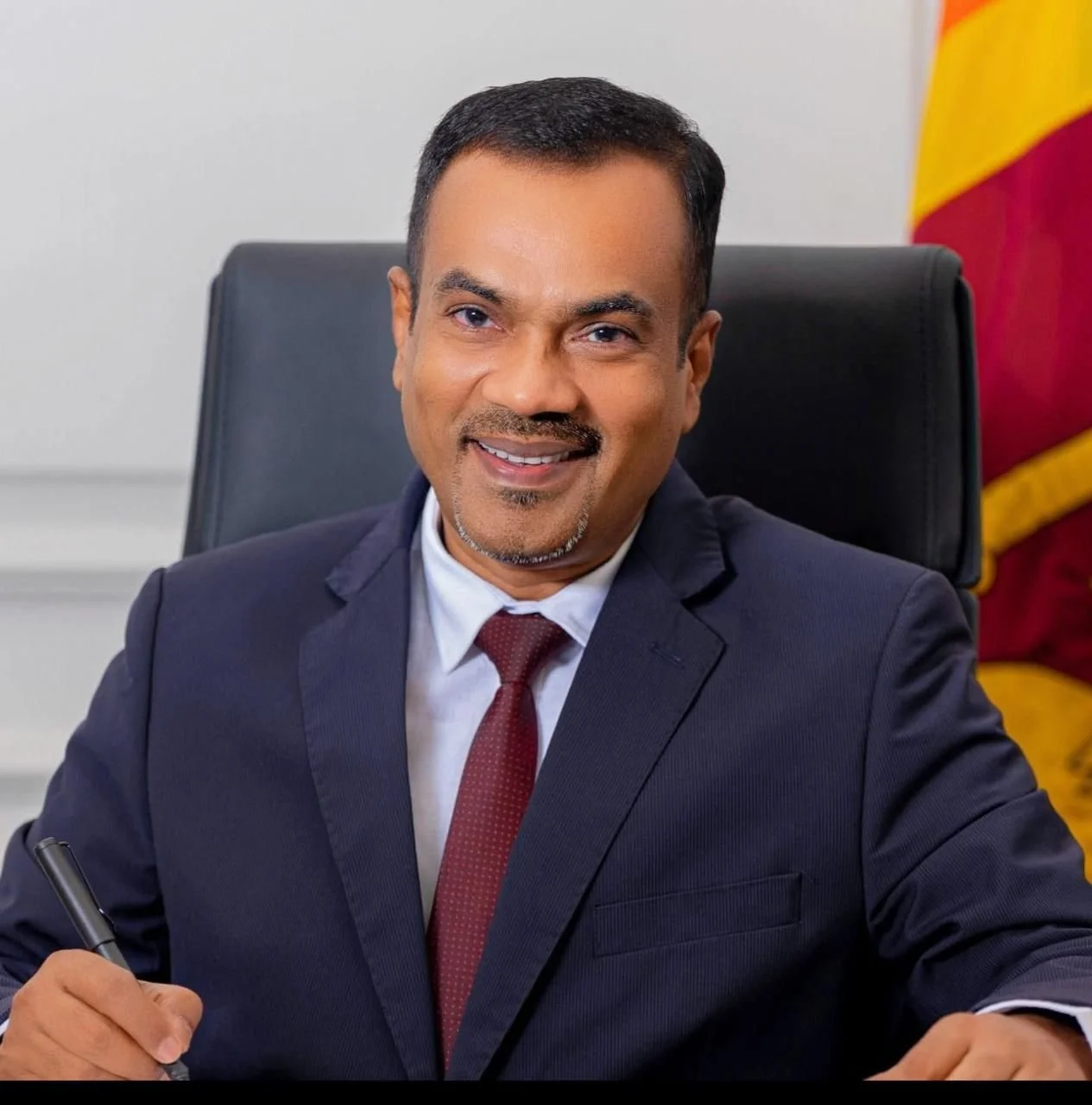 A man in a suit sitting at a desk, smiling, with a pen in his hand and a flag in the background.