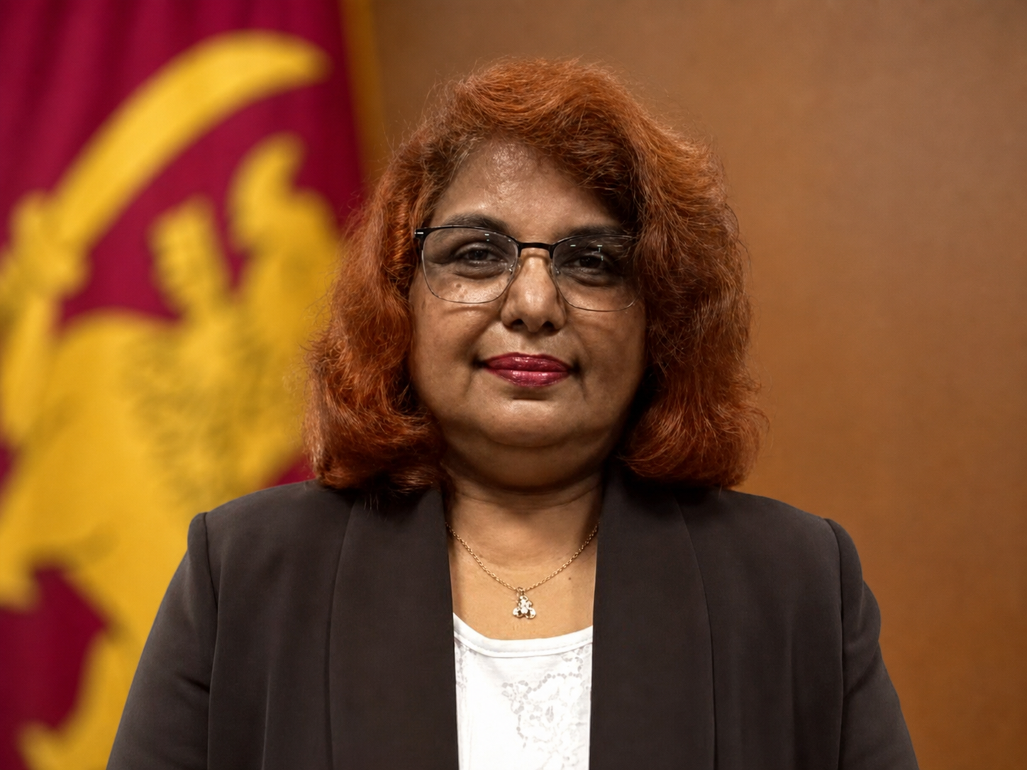 A woman with curly red hair and glasses wearing a black blazer and white top, standing in front of a yellow and red emblem or flag.