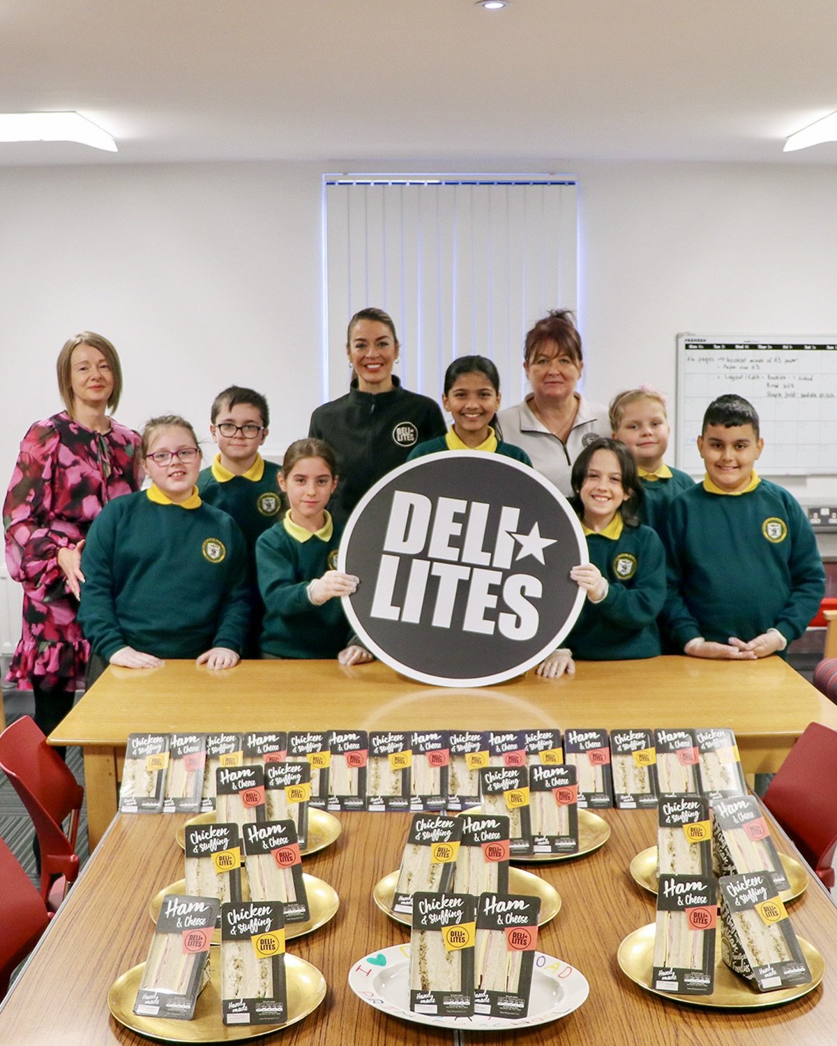 Group of children and adults standing behind a table with DELI LITES sandwiches, holding a sign that reads "DELI LITES," with packaging of chicken and ham sandwiches for DELI LITTLES scheme.
