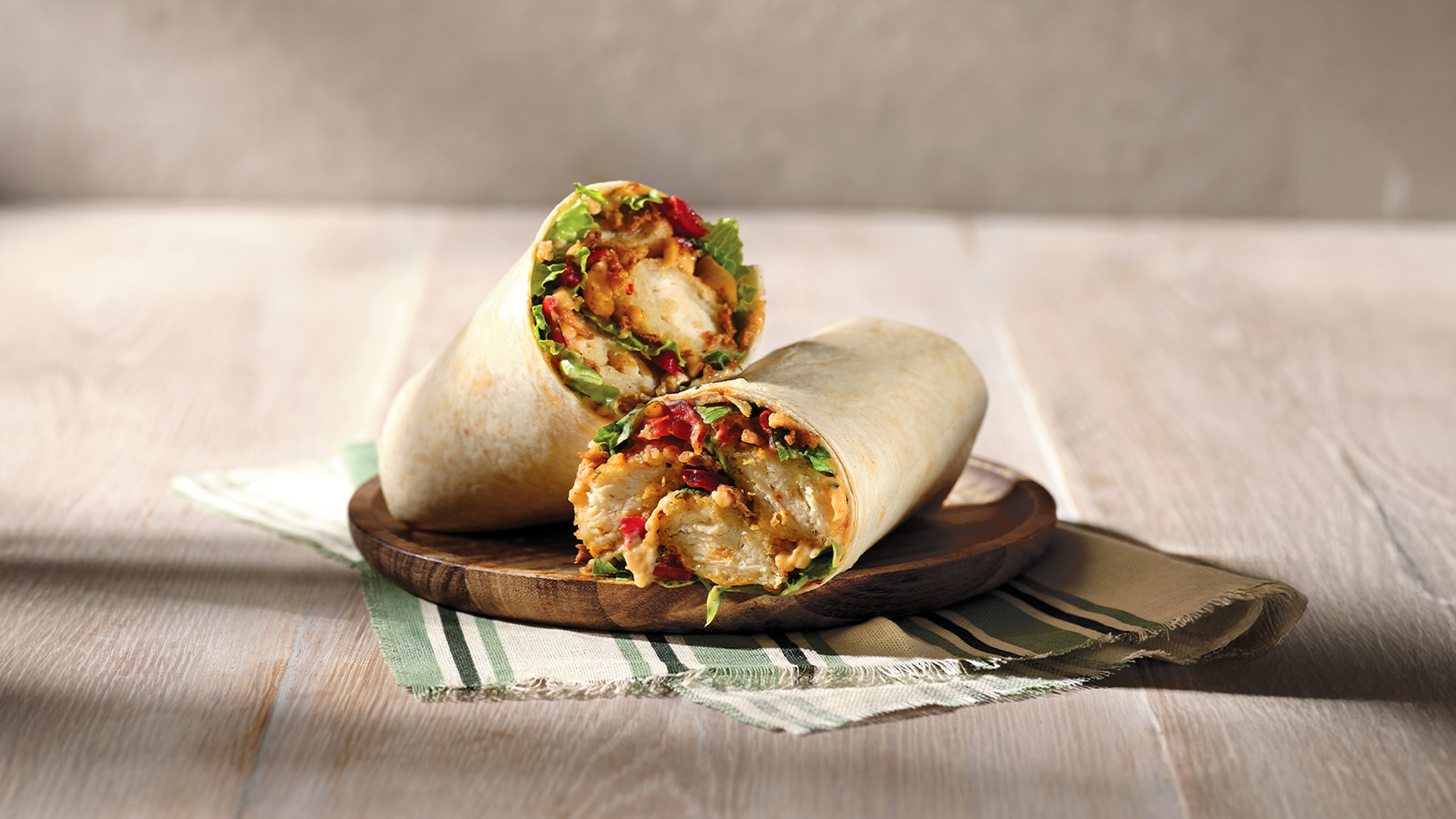 Two chicken wraps with lettuce, red peppers, and sauce on a wooden plate, placed on a striped cloth on a wooden table.